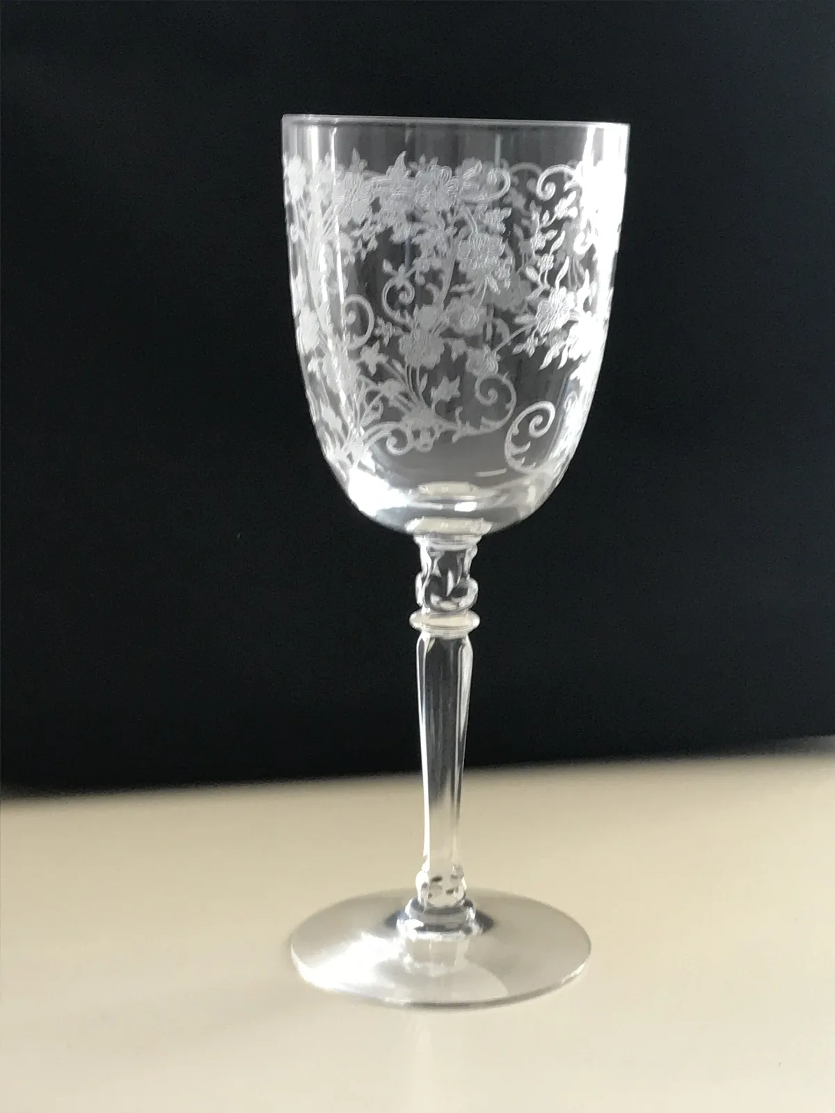 Clear stemmed goblet against a black background, the bowl covered with a fine white floral etching, the stem faceted with a small knop above the foot