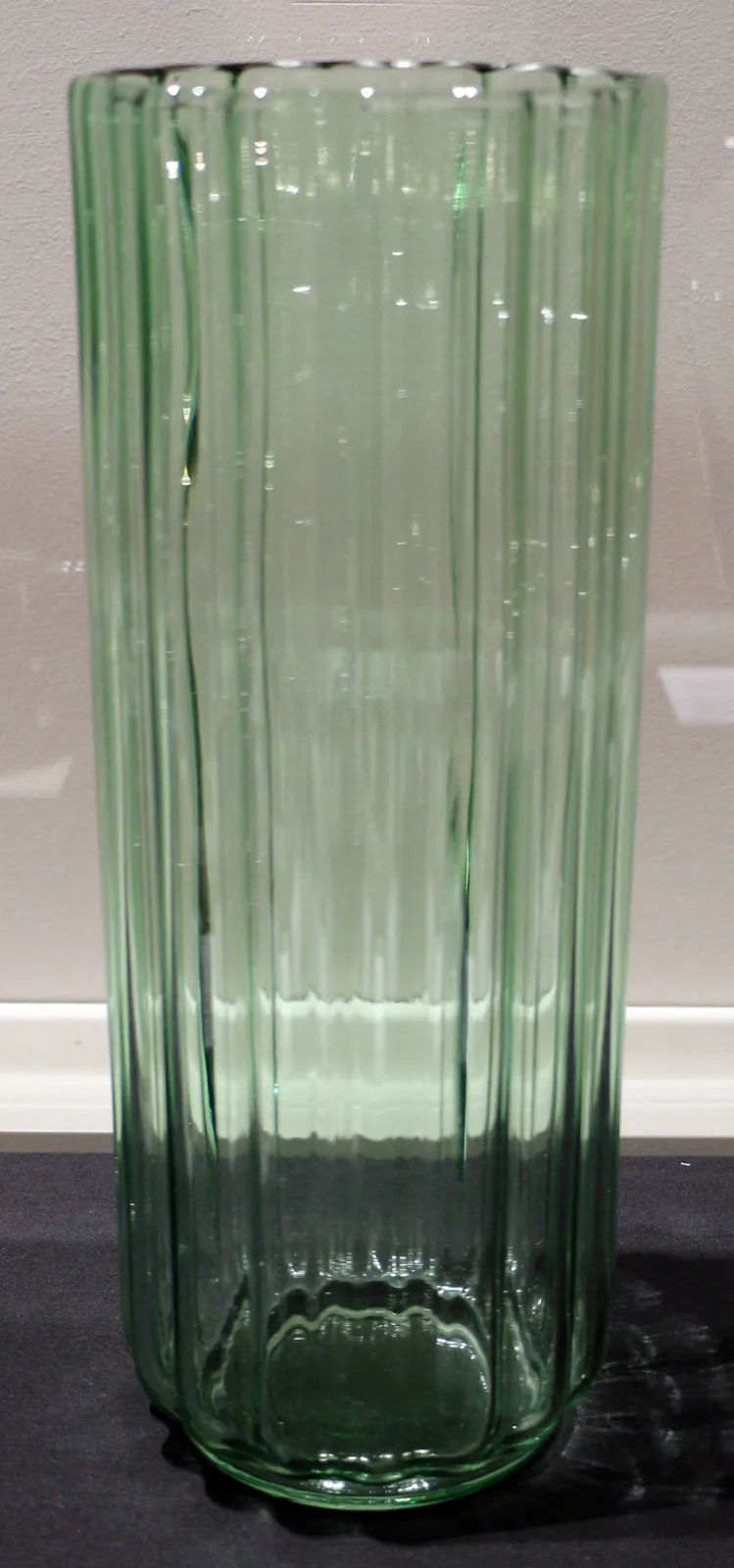 Tall cylindrical vase of pale green glass with deep vertical ribs running the full height, on a plain background