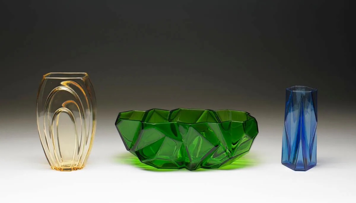 Three pieces of Art Deco glass on a graduated grey background: an amber ribbed vase at the left, a green angular faceted bowl at the centre, and a blue hexagonal vase at the right