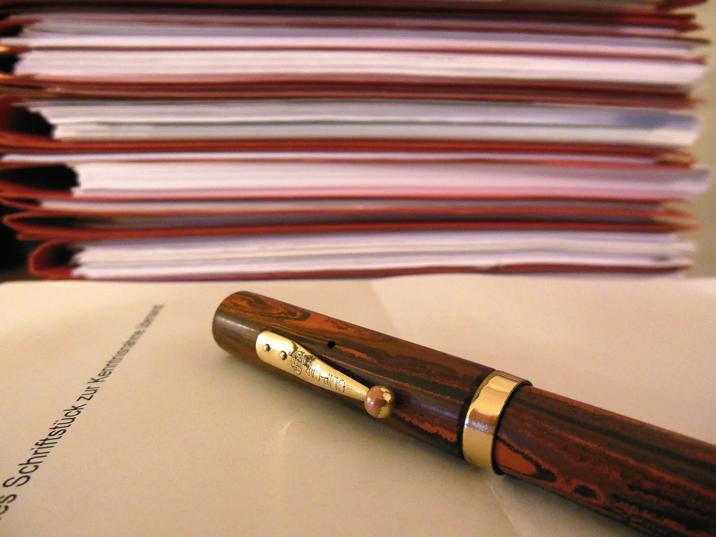 Slim capped fountain pen in mottled red-brown and black material with a narrow gold band at the cap lip and a gold clip, lying at an angle on a sheet of paper