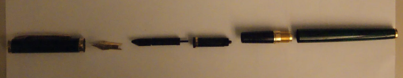 A fountain pen dismantled into six parts laid in a row on a pale surface: the cap, the barrel, the section with its nib and feed, a rubber sac, and two smaller fittings