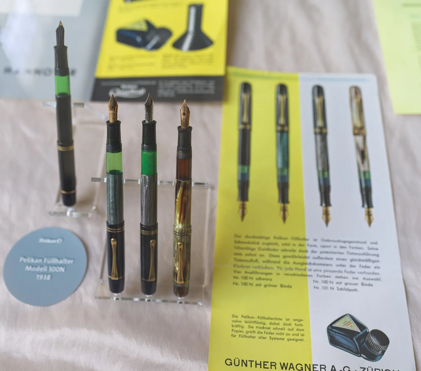 Four green-striped fountain pens with gold nibs displayed upright in a case beside an original printed leaflet in German describing the model