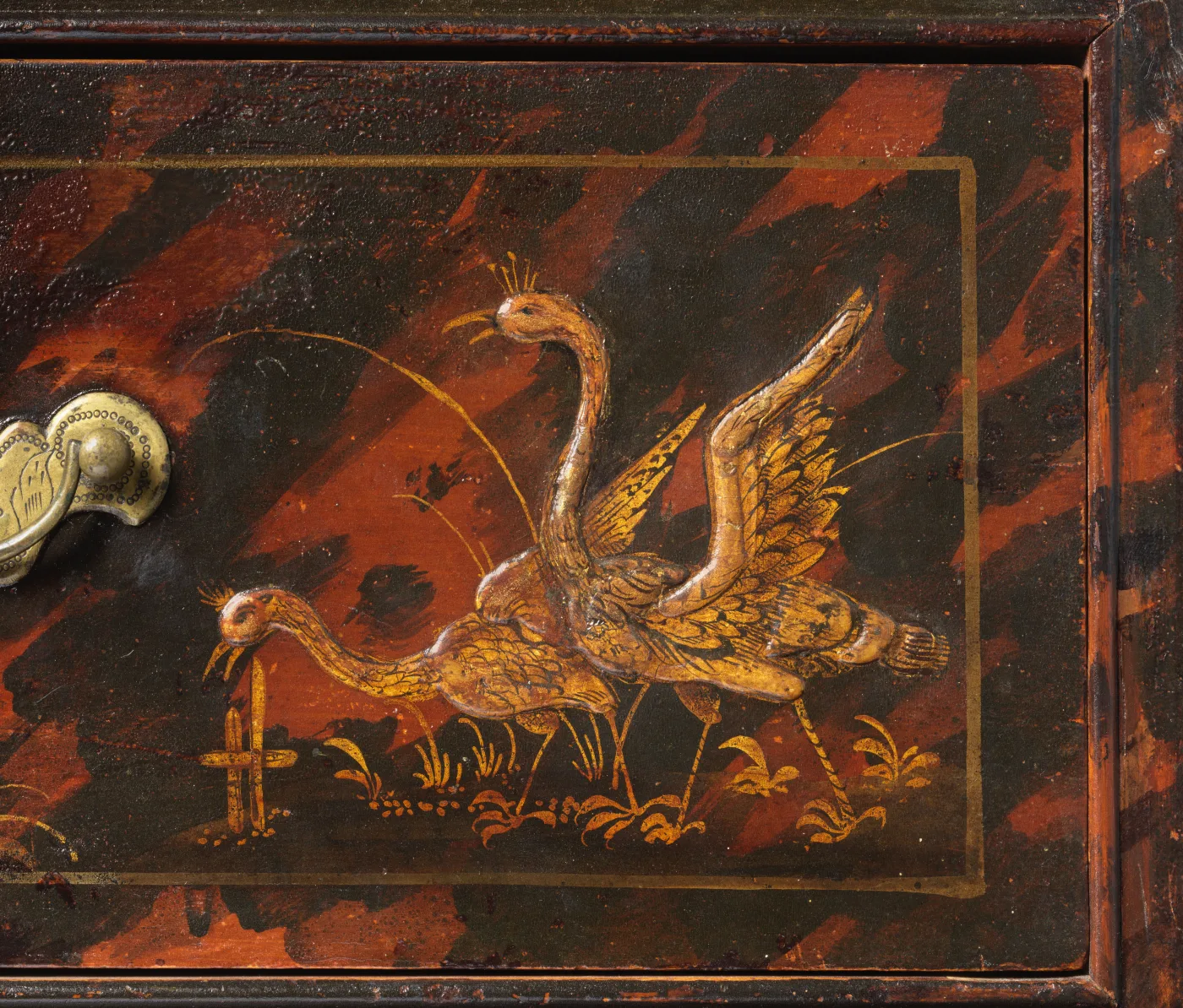 Close view of a japanned drawer front showing two gilt swans in raised relief over a mottled red and black ground, with a brass pull at the edge
