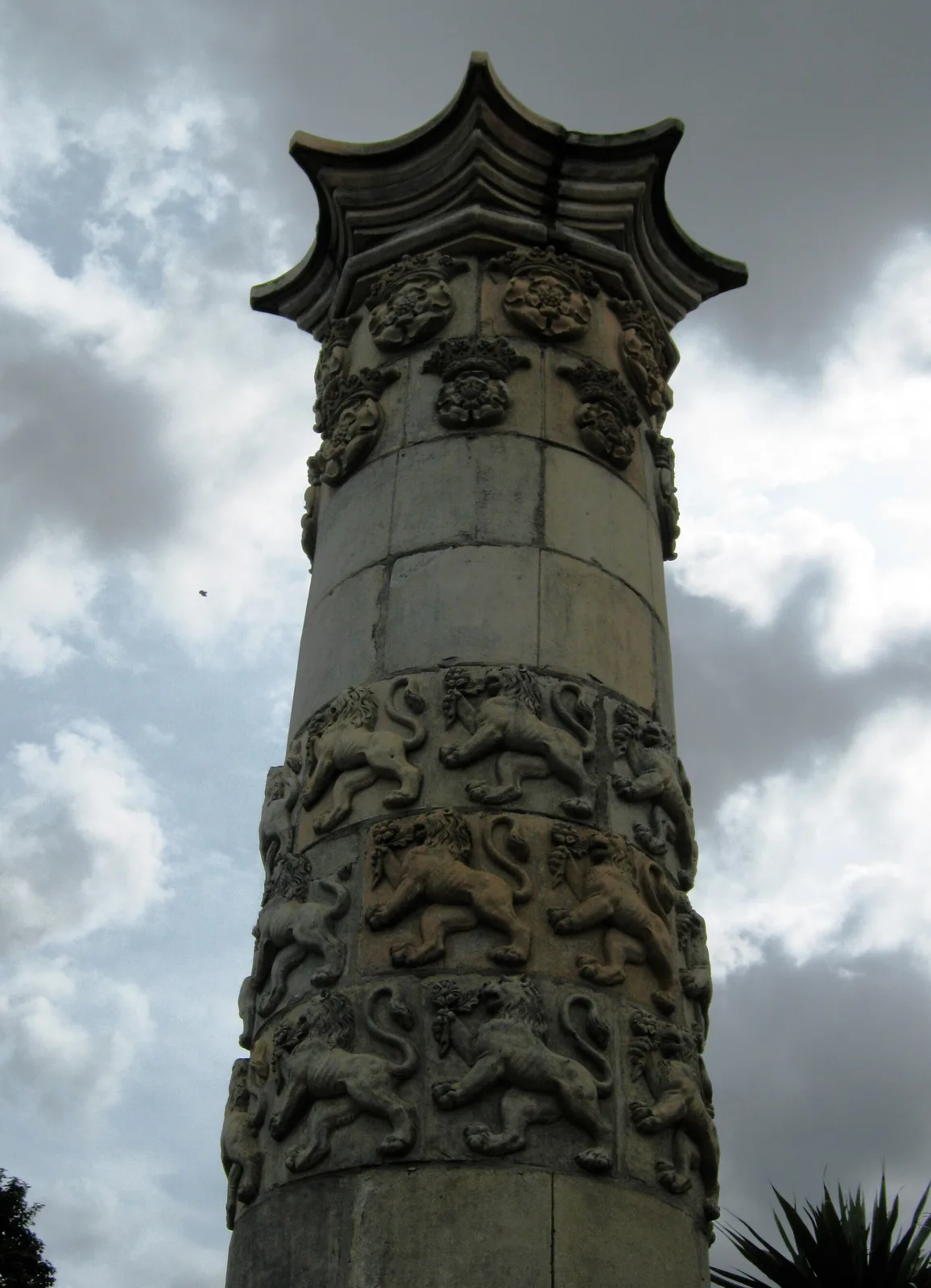 Tall pale stone pillar photographed against cloud, built up from curved blocks and banded with rows of identical moulded lions and crowned Tudor roses