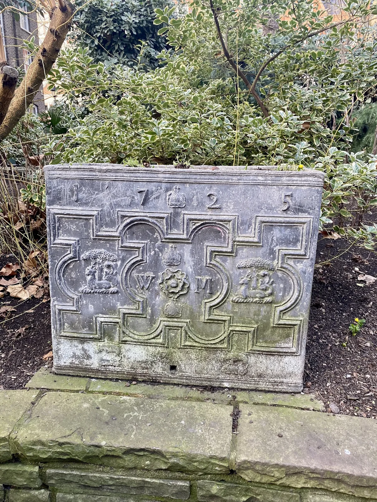 Rectangular lead water cistern standing in a shrub bed, its front cast in relief with strapwork panels, a Tudor rose flanked by the initials W and M, and the date 1725 spread across the top rail around a crown