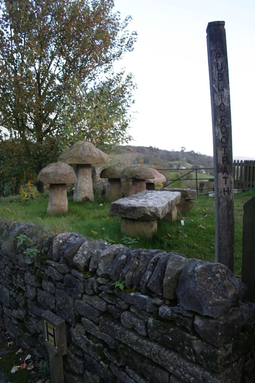 Five mushroom-shaped stones and a slab bench arranged on grass behind a dry stone wall beside a public footpath sign, one carrying a small sale label