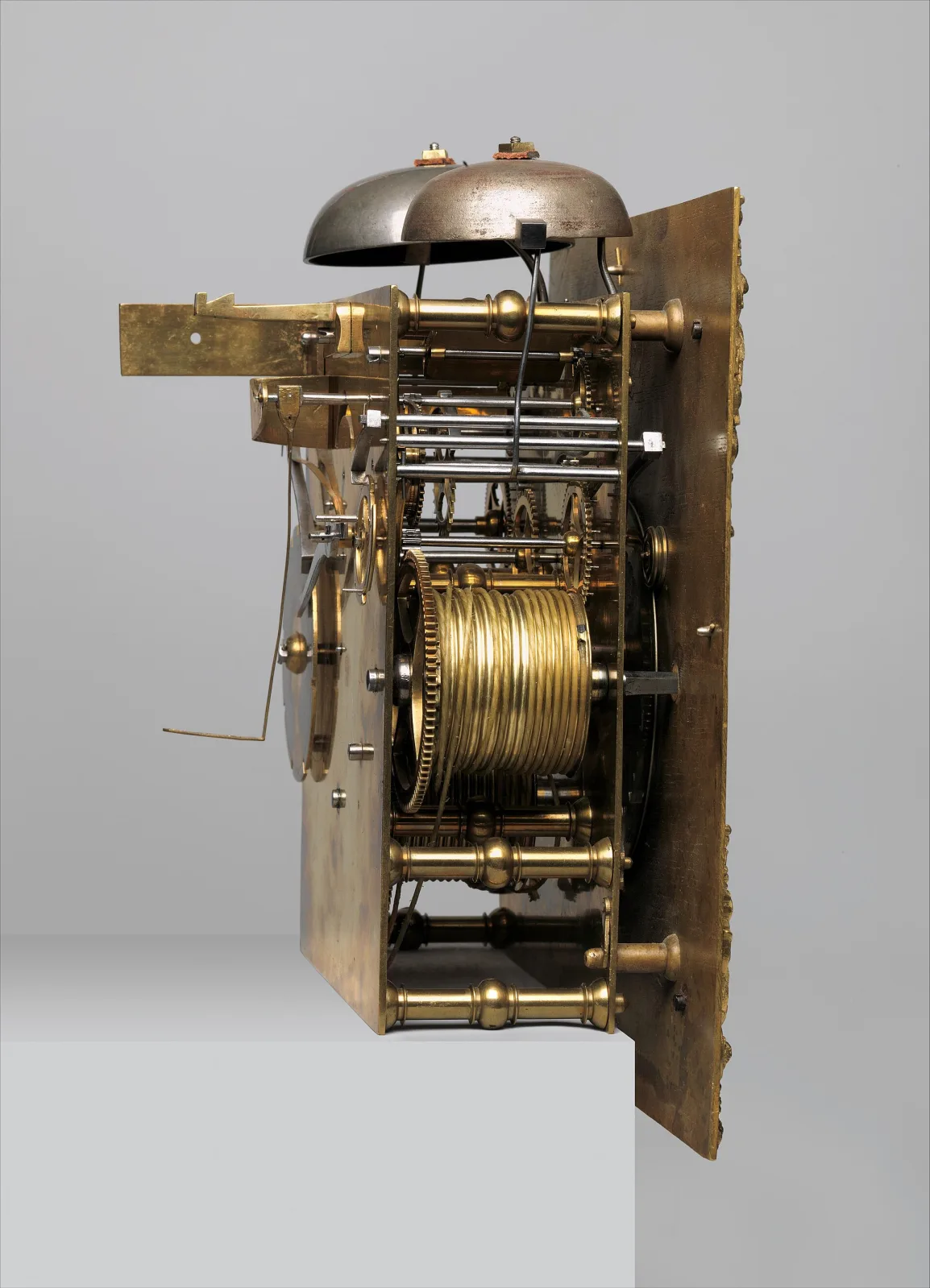 Clock movement removed from its case, seen from the side: two brass plates held apart by pillars, carrying a train of toothed wheels, with a large bell mounted above and a hoop and pins at the top