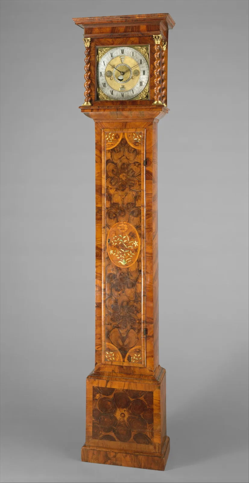 Tall narrow longcase clock veneered in figured walnut marquetry, with a flat-topped hood on turned columns, a rectangular brass dial and a slim trunk over a plain plinth