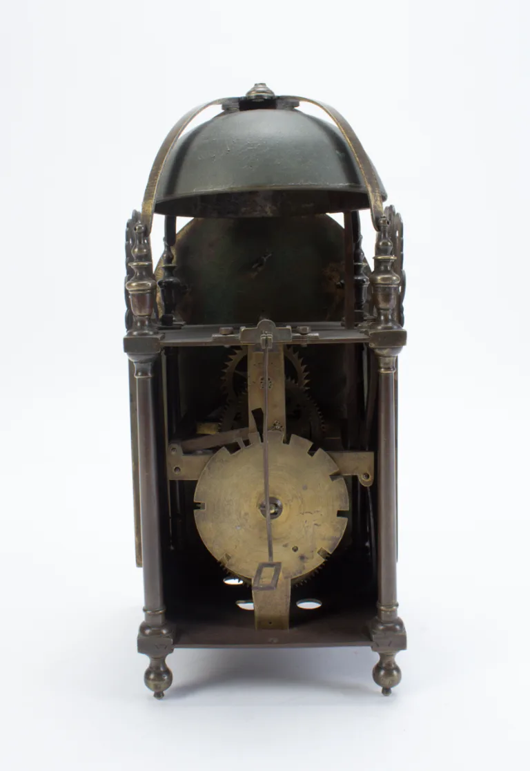Small brass lantern clock with its side door swung open, showing the vertical arrangement of wheels and the bell frame above, standing on four turned feet against a pale ground