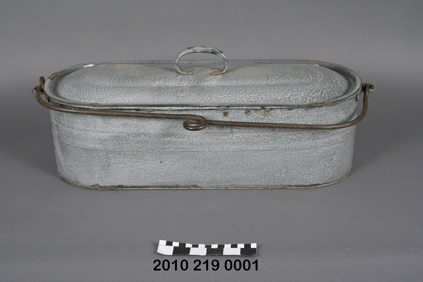 Long oval covered vessel in pale grey mottled enamel with a wire bail handle and a small enamelled loop on the lid, chipped to bare rusting metal at rim and base