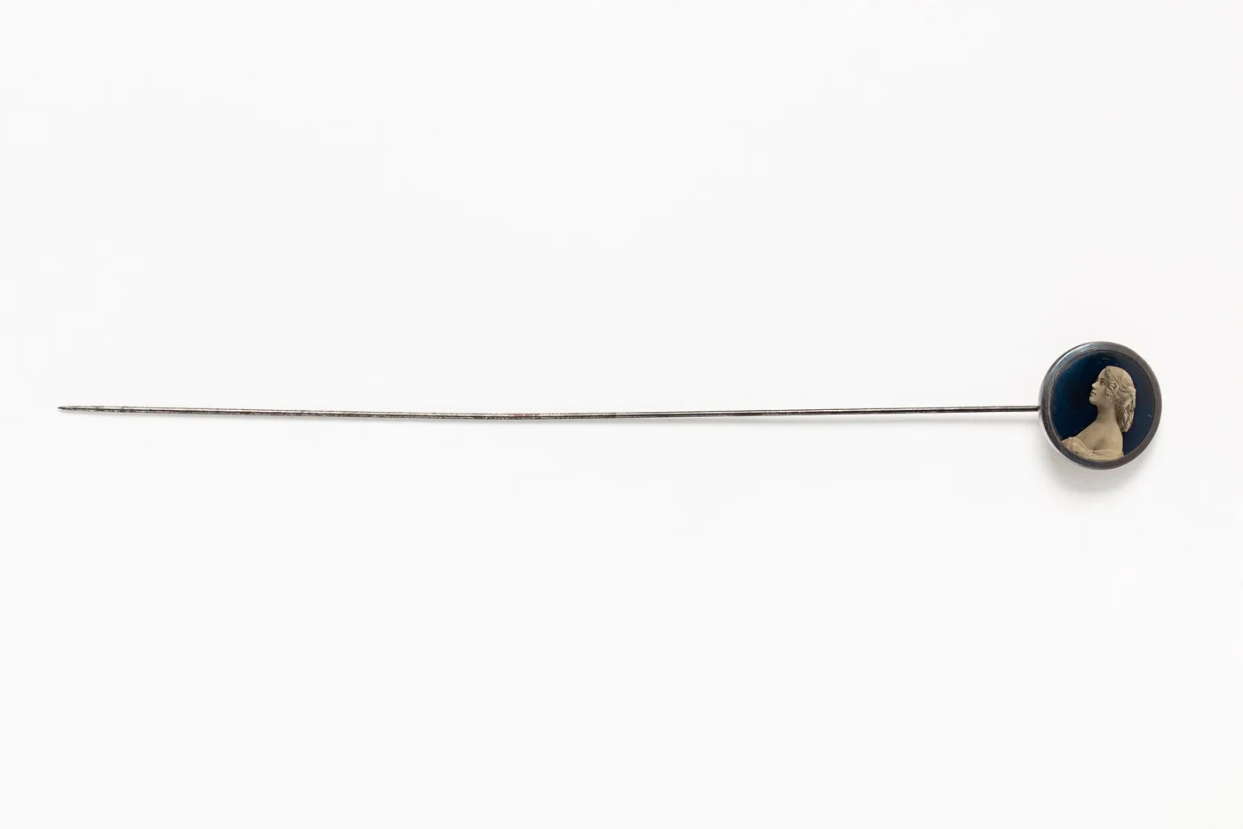 Long steel hat pin lying flat, with a round dark blue head carrying a pale profile bust of a woman in relief