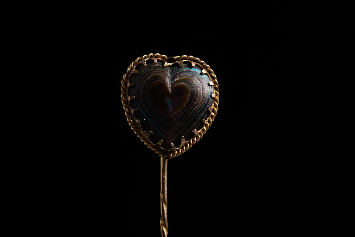 Heart-shaped hat pin head cut from iridescent abalone shell, held by claws inside a twisted gold-coloured rope border, on a twisted shaft