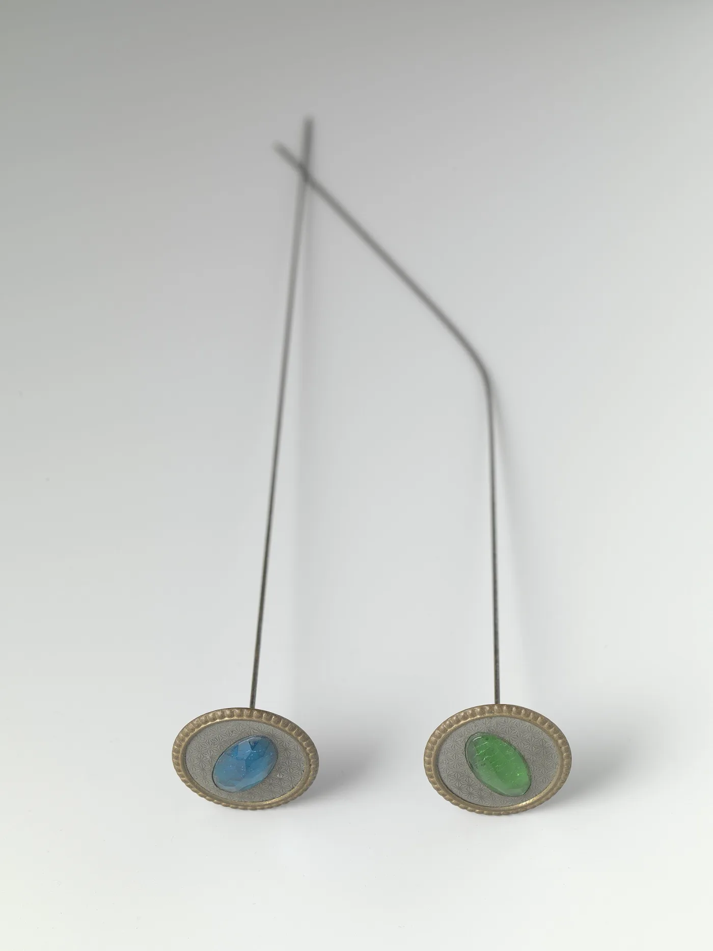 Two hat pins with oval gilt-rimmed heads set with faceted blue and green glass stones over an engine-turned silver ground; the right-hand shaft is bent at a sharp angle near the top