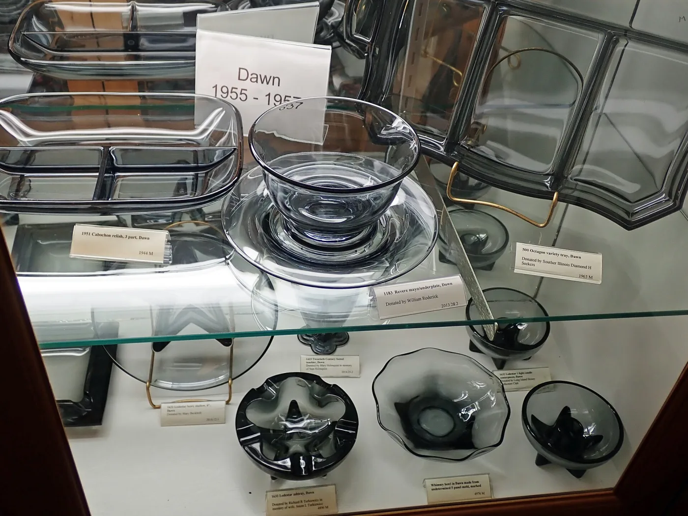 Museum case of smoky grey glass trays, bowls and ashtrays behind a card reading Dawn 1955 to 1957