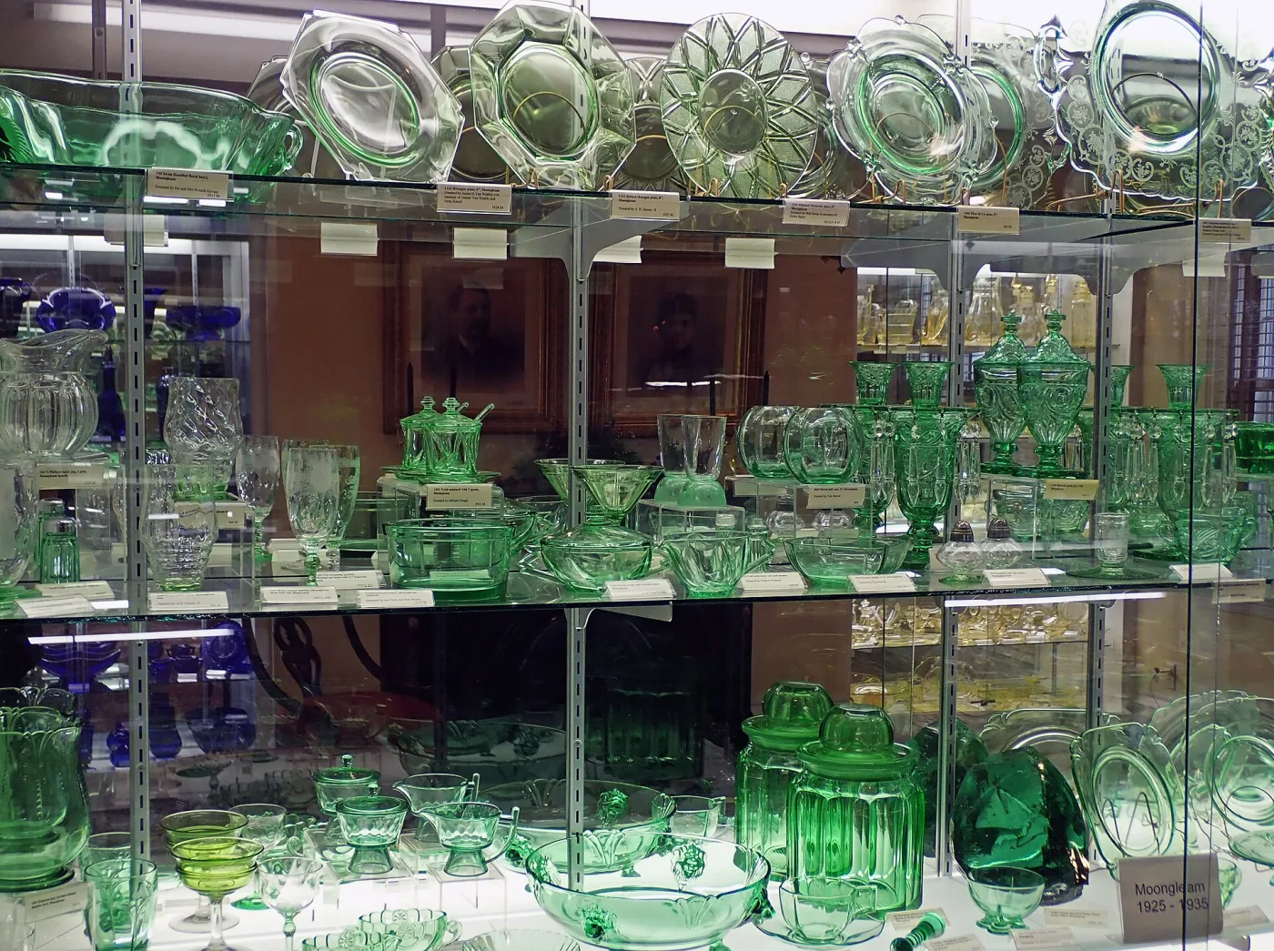 Museum display case filled with green Heisey glass &mdash; stemware, plates, bowls, candlesticks and a cornucopia vase &mdash; arranged on glass shelves