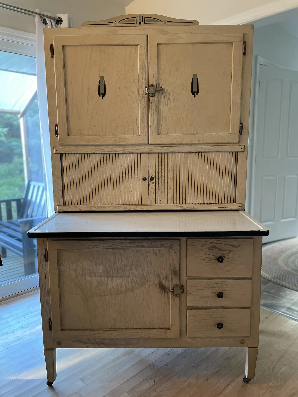 Kitchen cabinet with the tambour closed across the middle of the upper case, showing a band of narrow vertical slats, stepped nickel escutcheons on the upper doors, a pierced stepped crest on top and a white enamel work surface with a black edge
