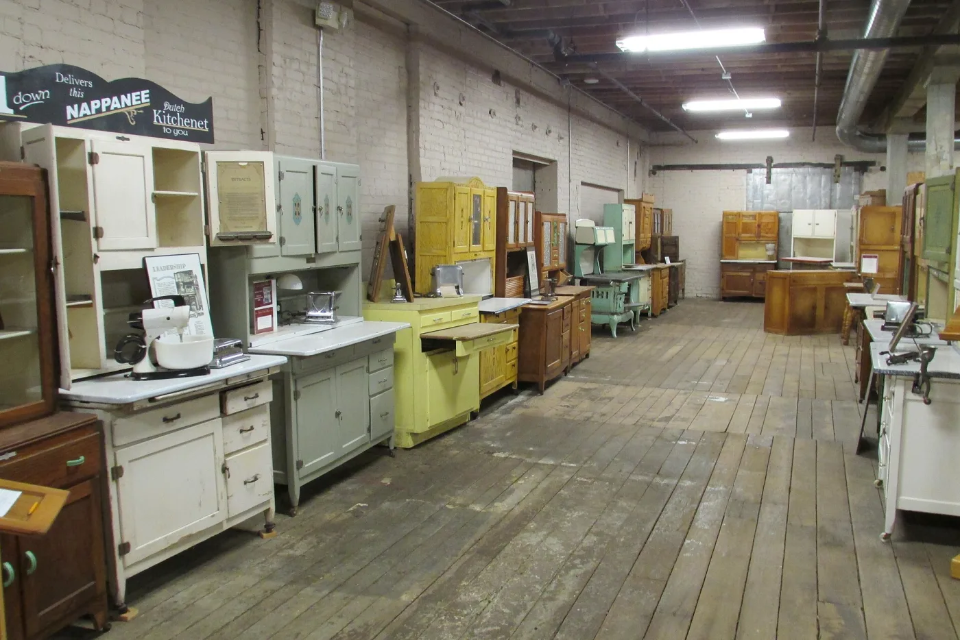 Long museum gallery lined with freestanding kitchen cabinets in white, grey-green, yellow, mint and golden oak finishes, with a painted dealer sign for the Nappanee Dutch Kitchenet hanging above them