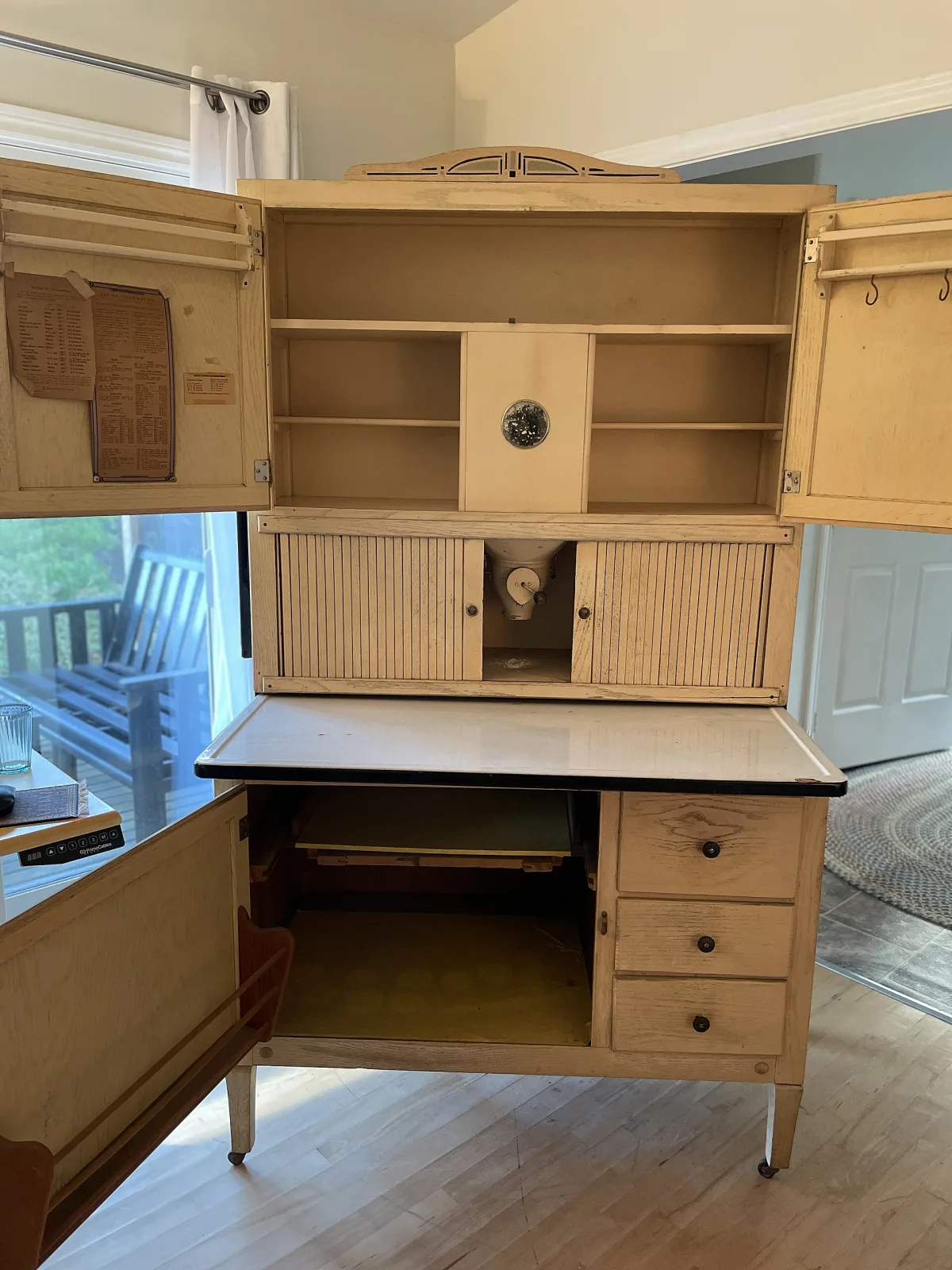 Kitchen cabinet fully open, showing a printed information chart pasted inside the left door, spice rails and S-hooks inside the right door, a flour bin front with a round glass sight window, the tambour slid apart to reveal a metal sifter cone and crank, and the lower case open on a sliding shelf