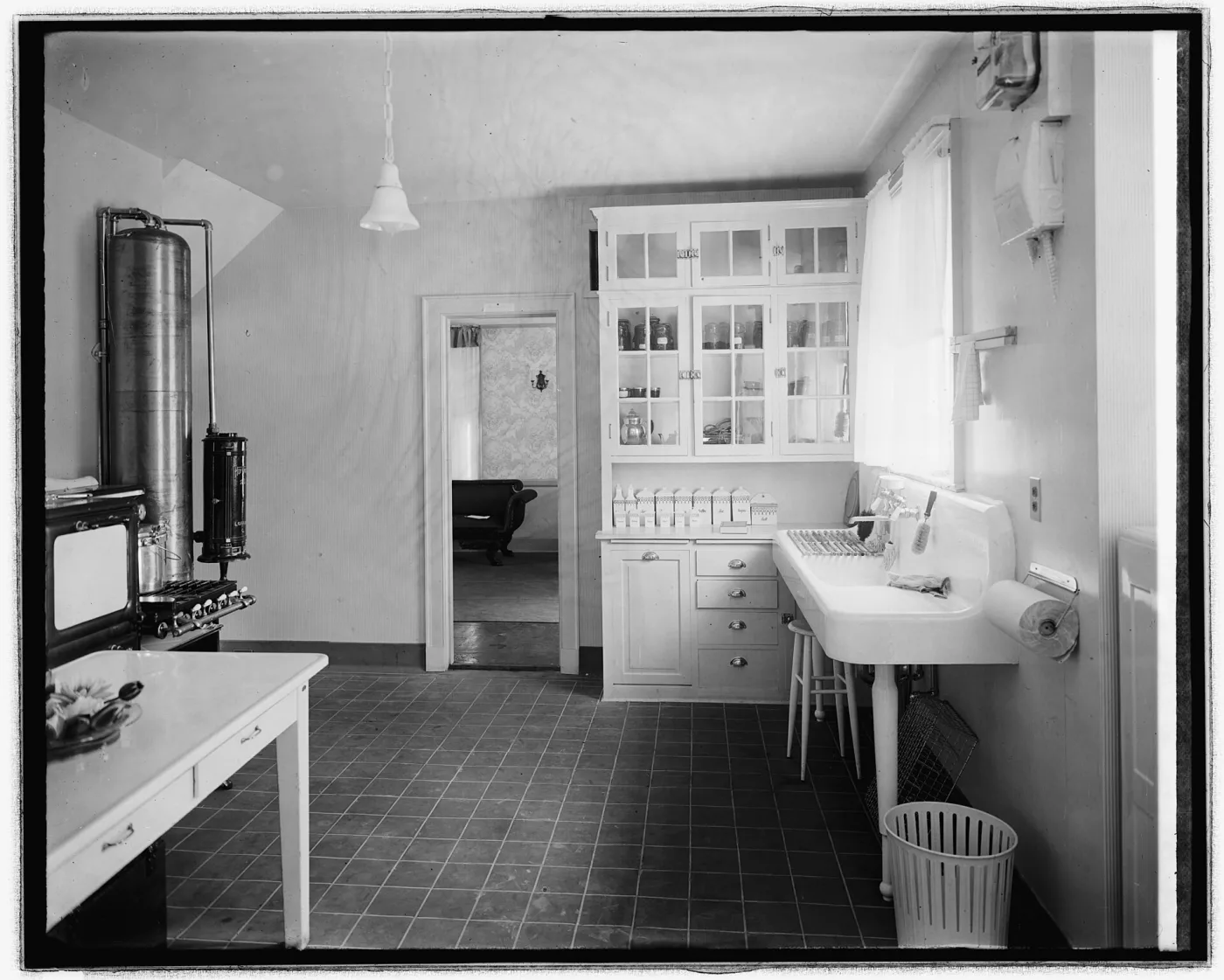 Black and white photograph of a white-painted kitchen around 1920, with a floor-to-ceiling built-in cupboard with glazed doors full of canning jars, a row of labelled canisters on the counter below, a tiled floor, an enamel table and a farmhouse sink