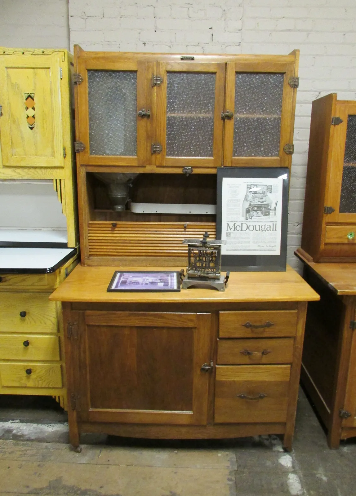 Golden oak kitchen cabinet with three upper doors glazed in heavily textured chipped glass, ornate dark cast hinges and latches, a small metal maker's plate on the top rail, and a galvanised flour bin with its sifter cone visible in the open centre compartment