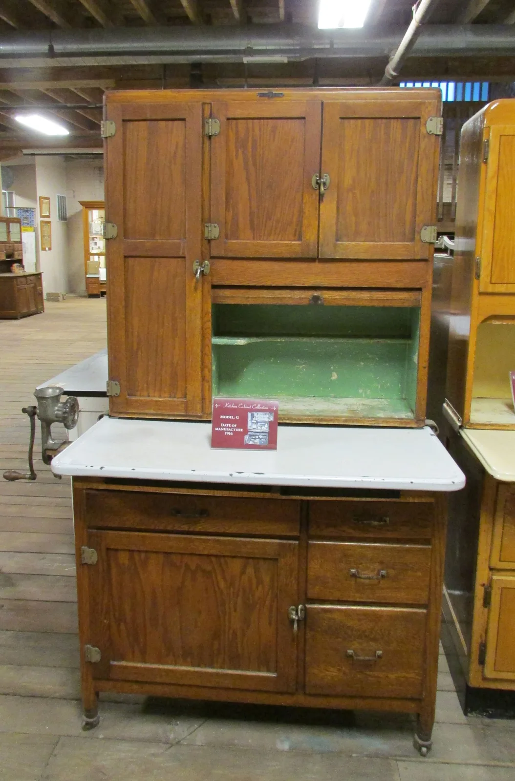 Golden oak kitchen cabinet with flat framed panel doors, brass butt hinges and latches, a green-painted open compartment, a white enamel pull-out work surface and a meat grinder clamped to its left edge