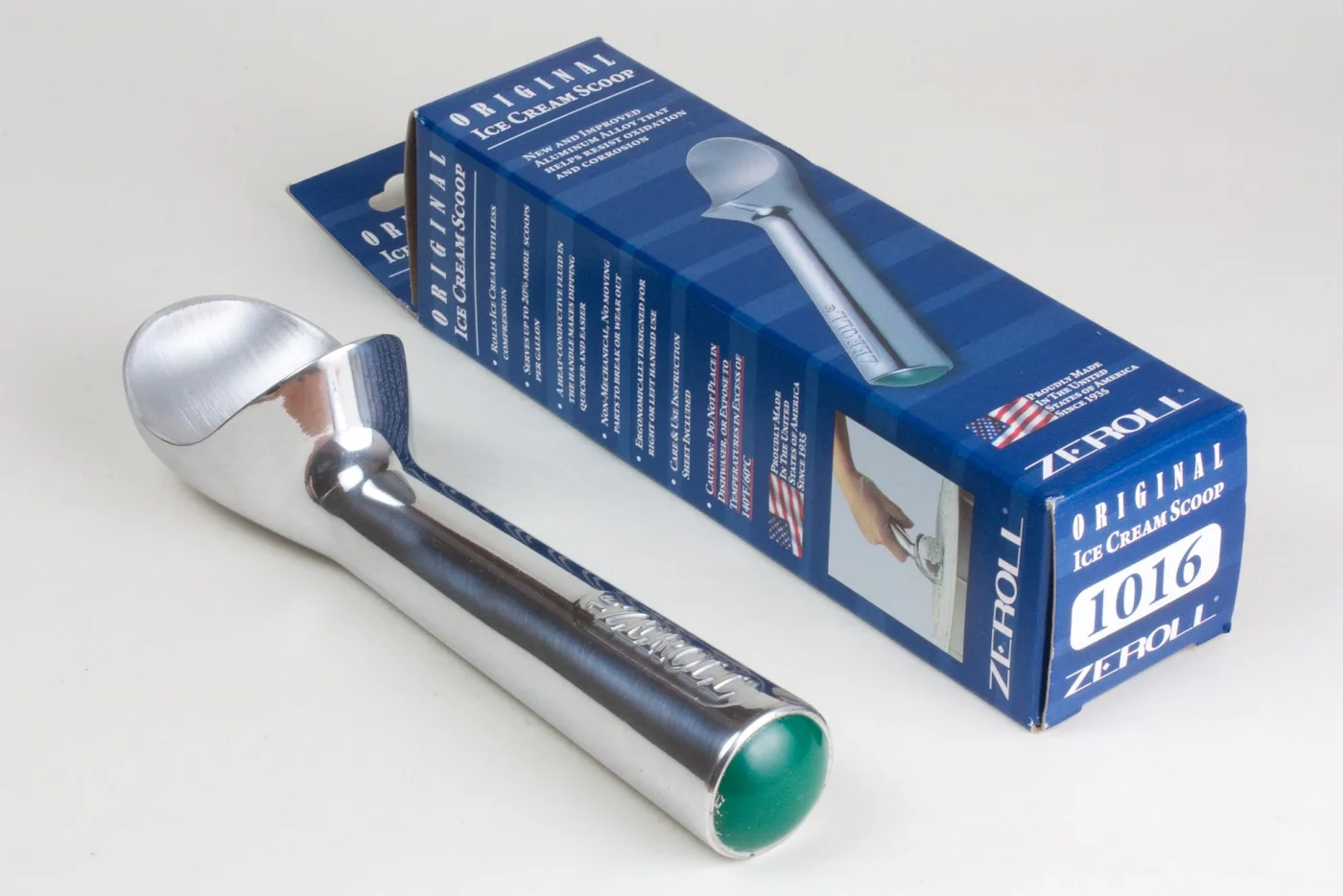 Polished one-piece aluminium Zeroll ice cream scoop lying beside its printed carton, with a coloured plug sealing the open end of the hollow handle