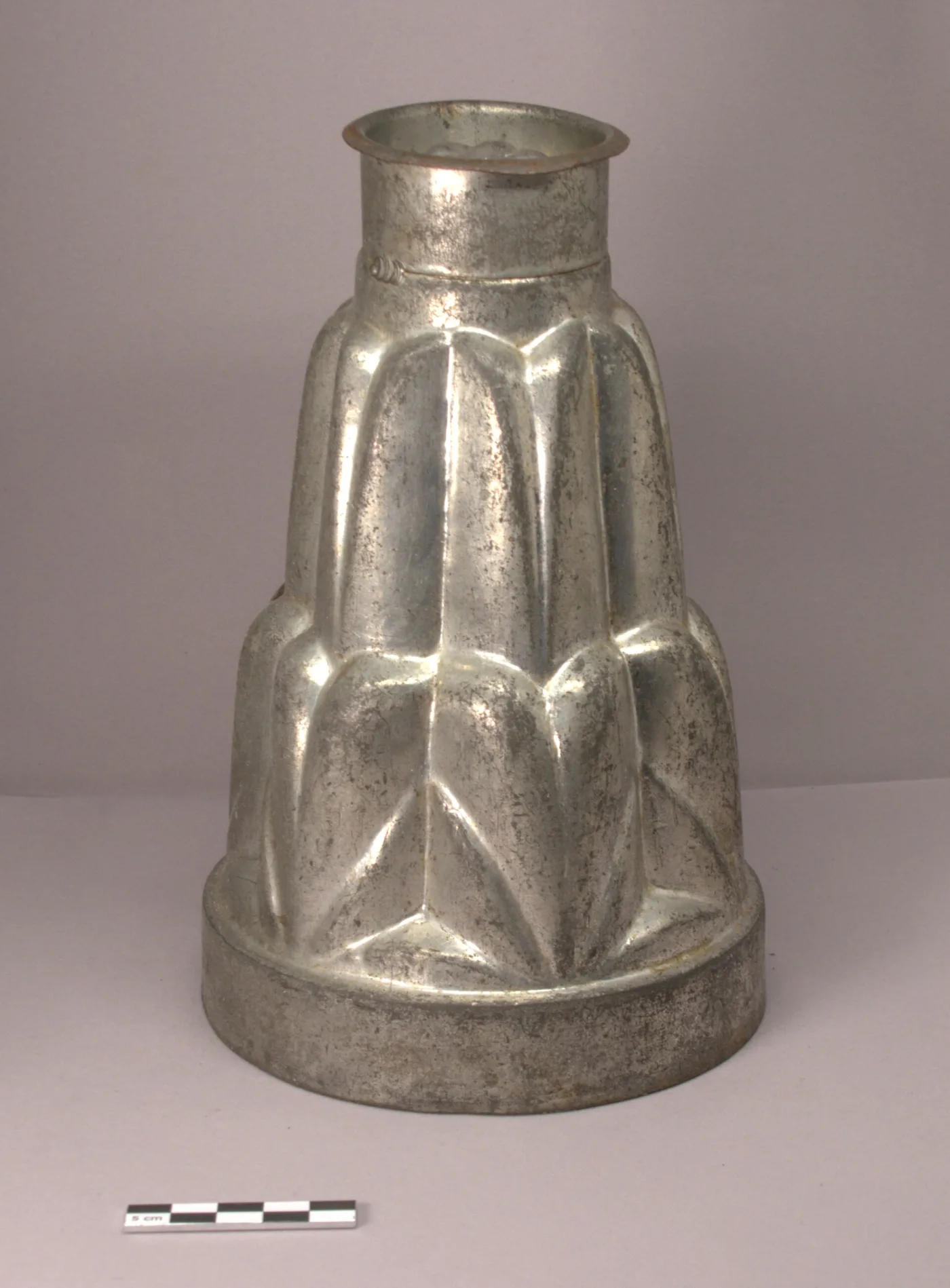Three-tier ice cream mould in tinned sheet iron, a stepped fluted column with a short collar at the top, standing beside a photographic scale