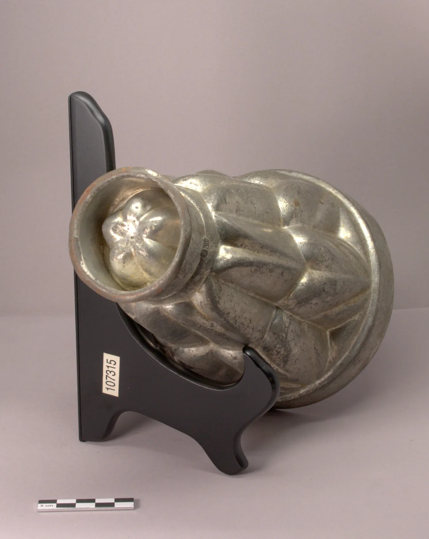 The tinned-iron tiered mould lying on its side on a display stand, its open collar showing the smooth fluted interior and a rosette at the closed end