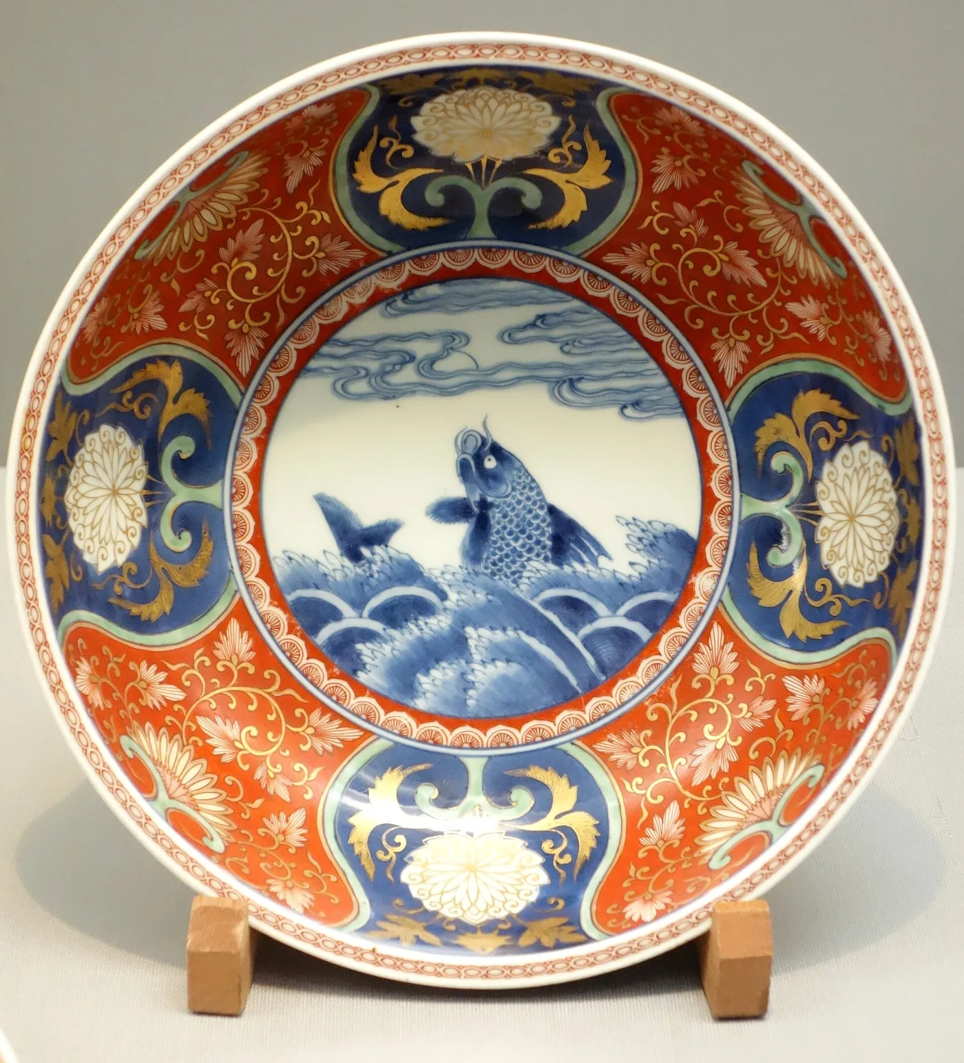 Wide porcelain bowl on a stand, its cavetto painted in underglaze blue with a fish among waves, the broad rim divided into panels of iron-red and gold scrollwork alternating with blue and white flowerheads