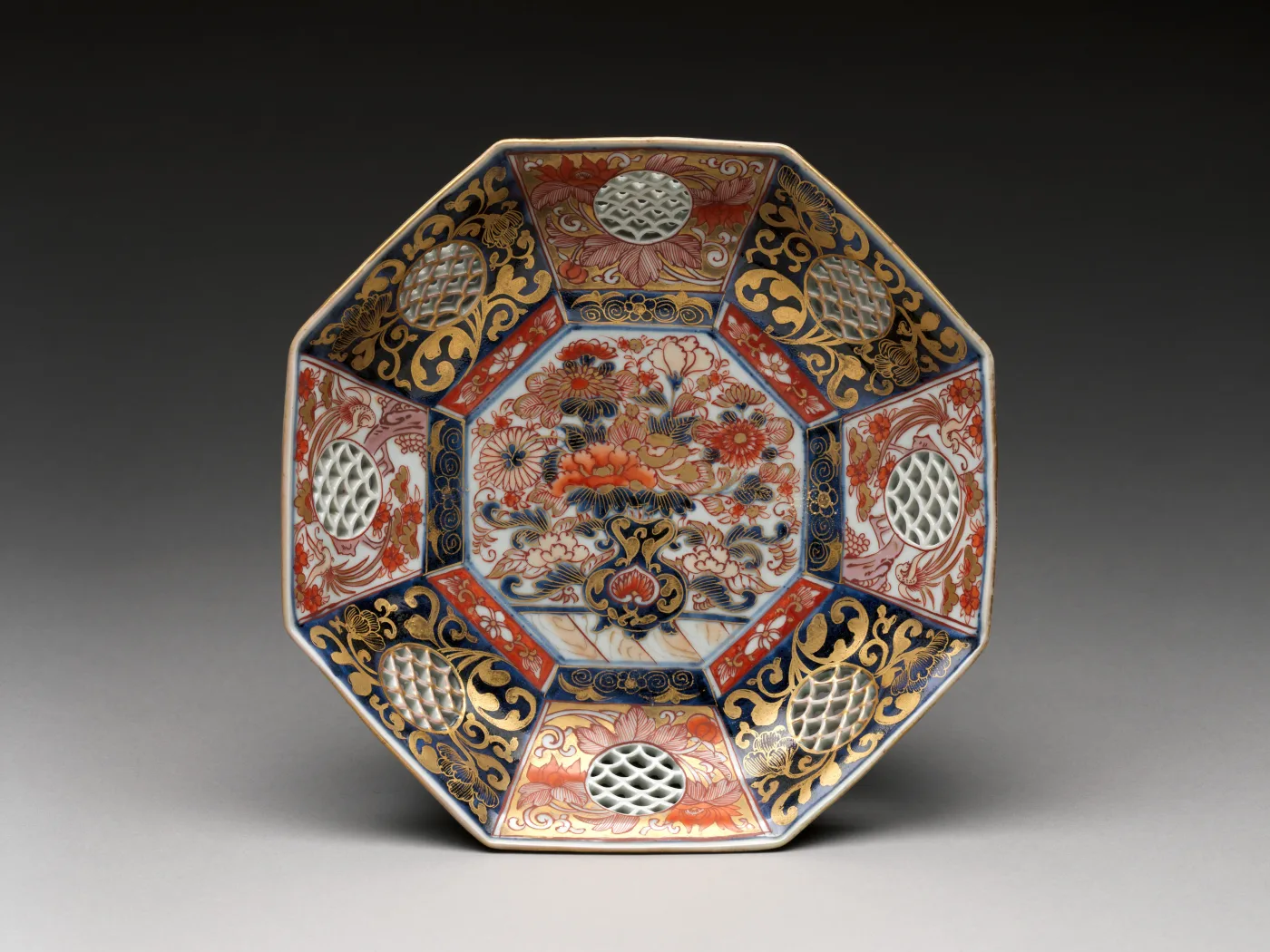 Octagonal porcelain dish densely covered in radiating panels of iron red, gold, blue and black, enclosing a central medallion with a vase of flowers, with no undecorated ground anywhere