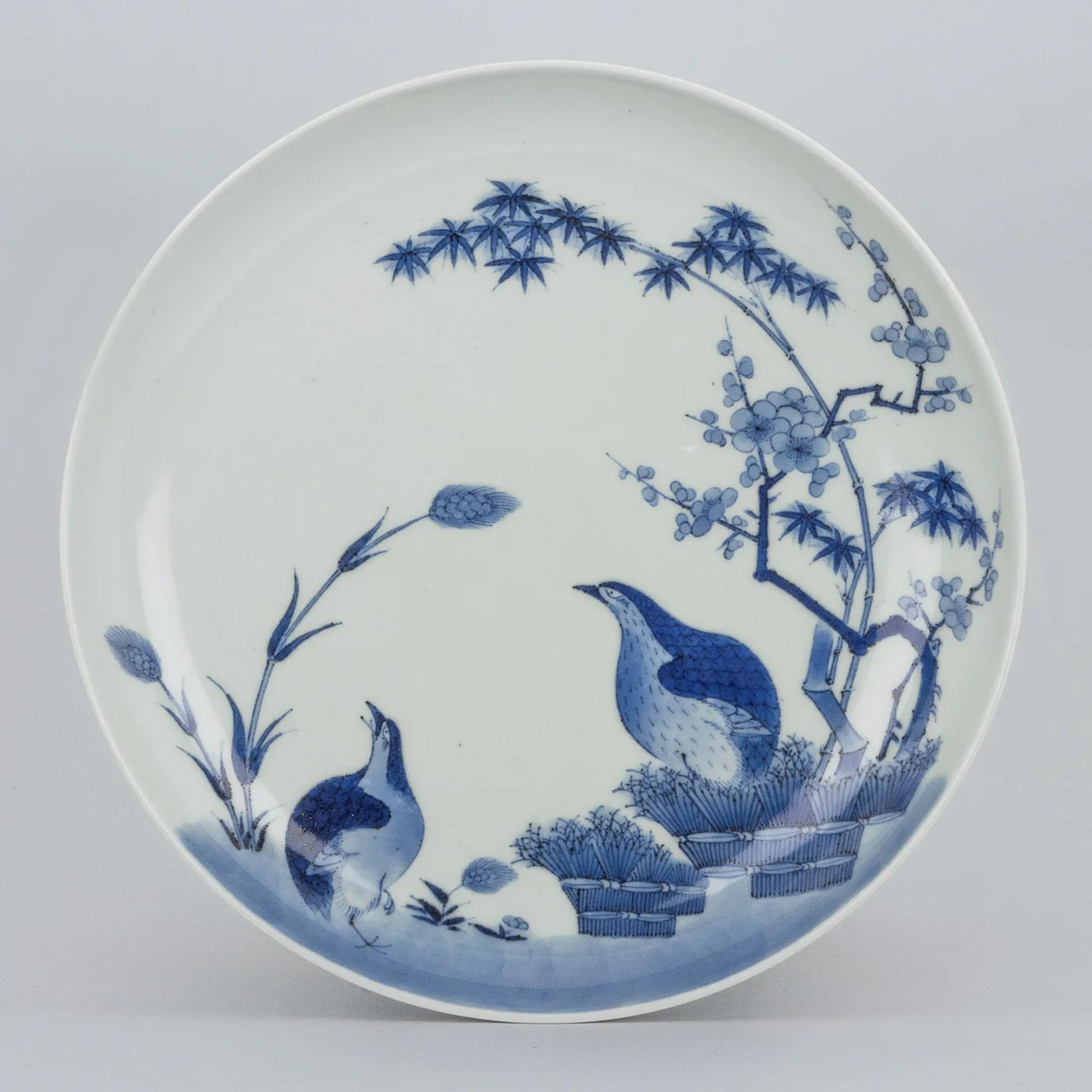 Shallow porcelain dish painted in underglaze blue alone, with a pair of quail among millet stems set off to one side of a wide plain white field