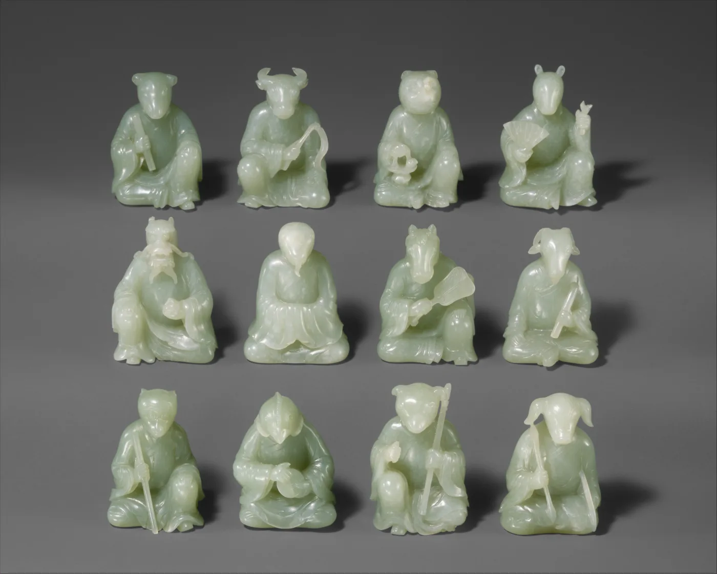 Twelve small seated figures with animal heads and human bodies, all carved in the same pale grey-green stone