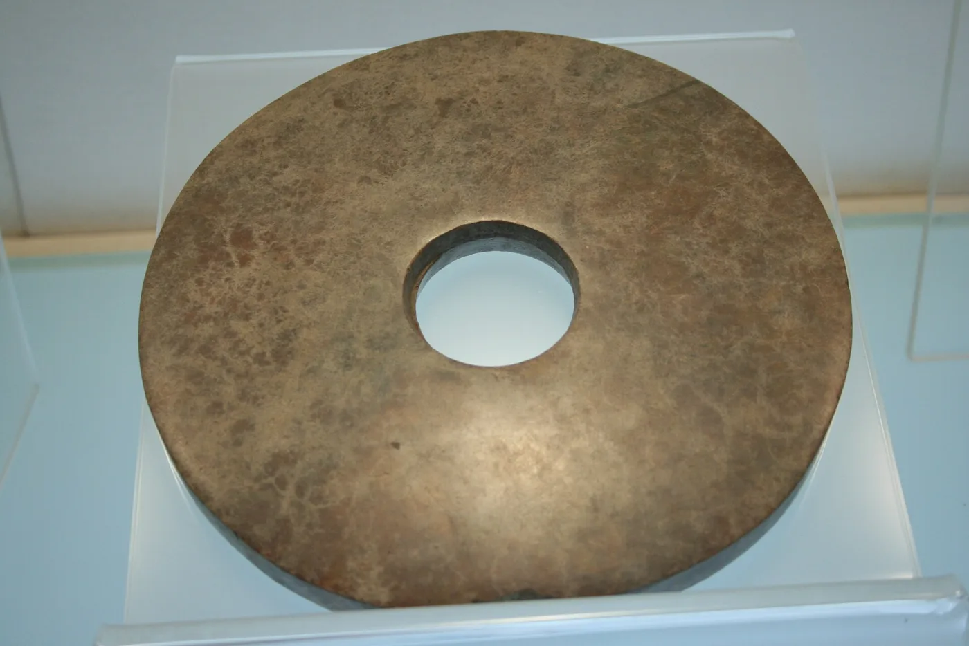 Large plain stone disc with a single central hole, mottled brown and grey, on a clear museum mount