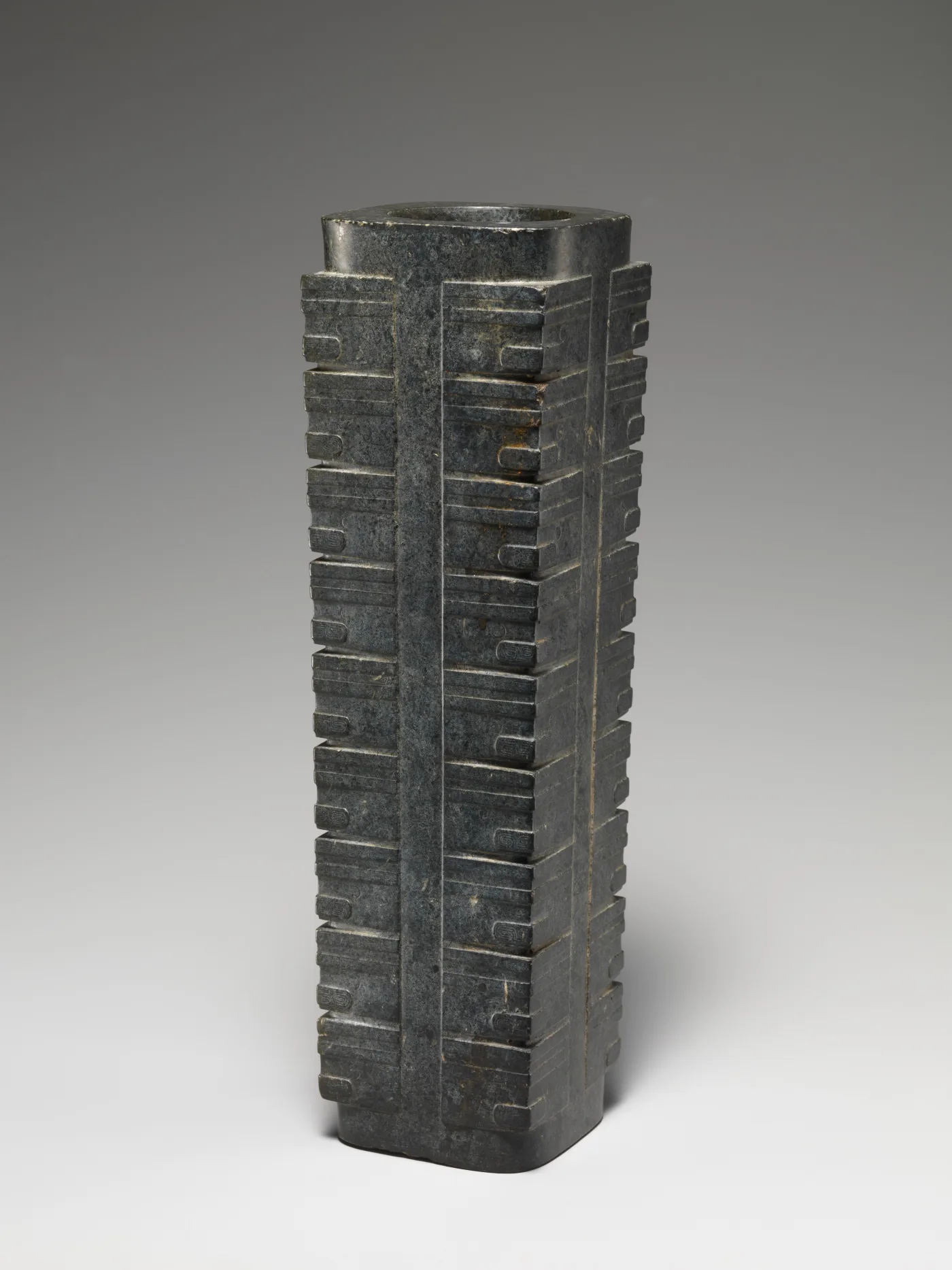 Tall squared tube of dark grey-green stone with a round bore, its four corners cut into stacked tiers of shallow bands