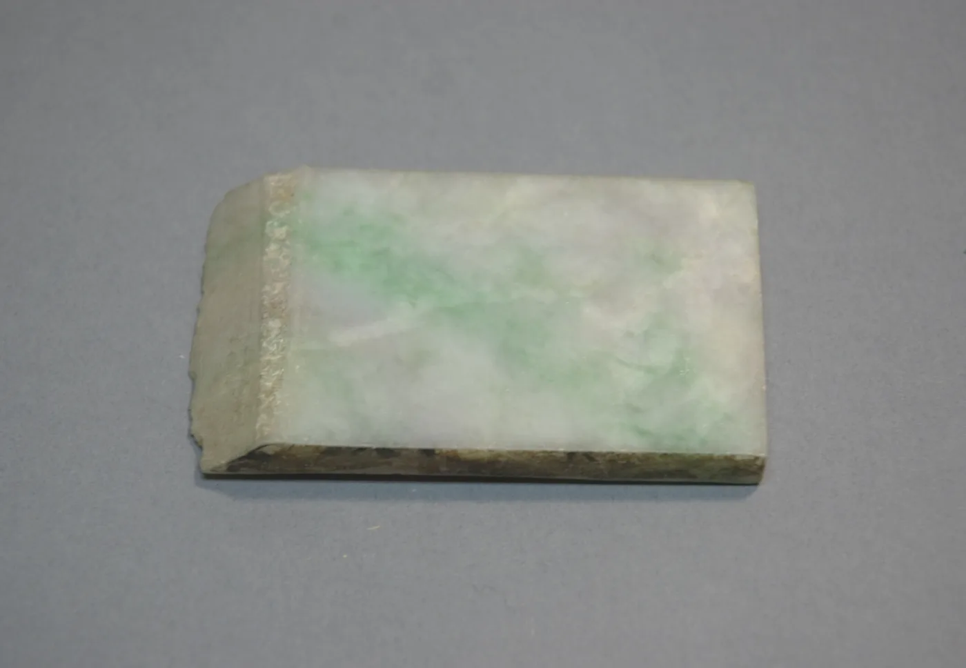 Sawn slab of pale jadeite, mottled white with drifting green patches and a rough brown weathered crust along one cut edge