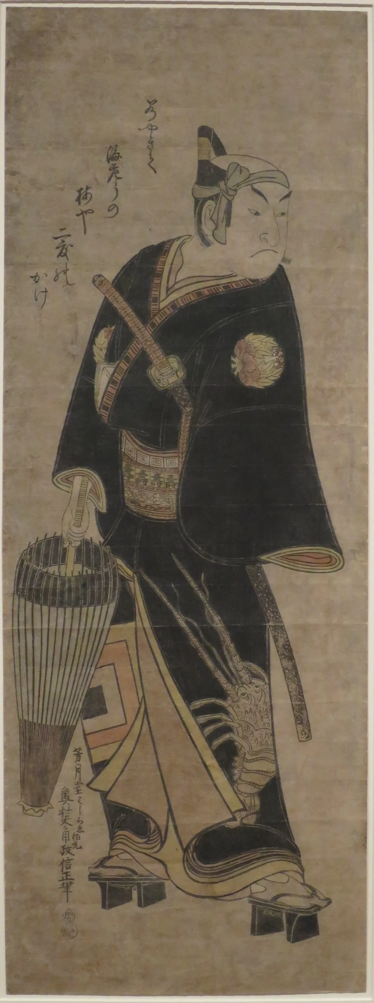 Tall narrow print of a kabuki actor in a black robe carrying a wicker lantern, with pale colour brushed on over black outlines