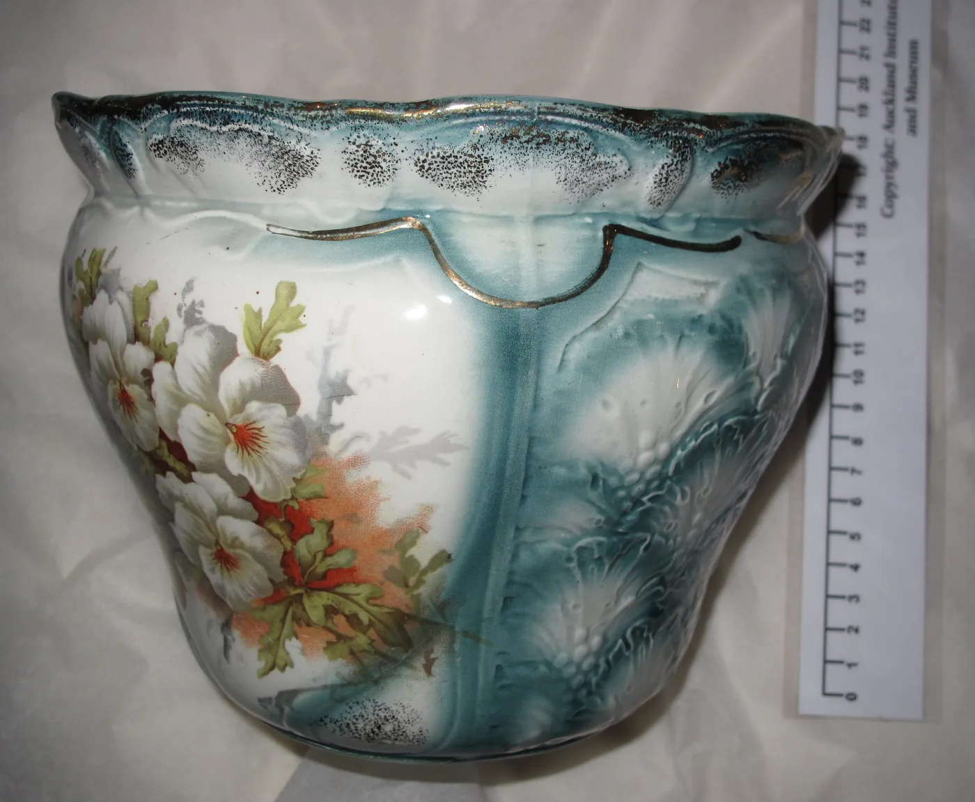 Ceramic jardiniere with white blossom and moulded leaf relief on a teal ground, photographed beside a paper scale rule