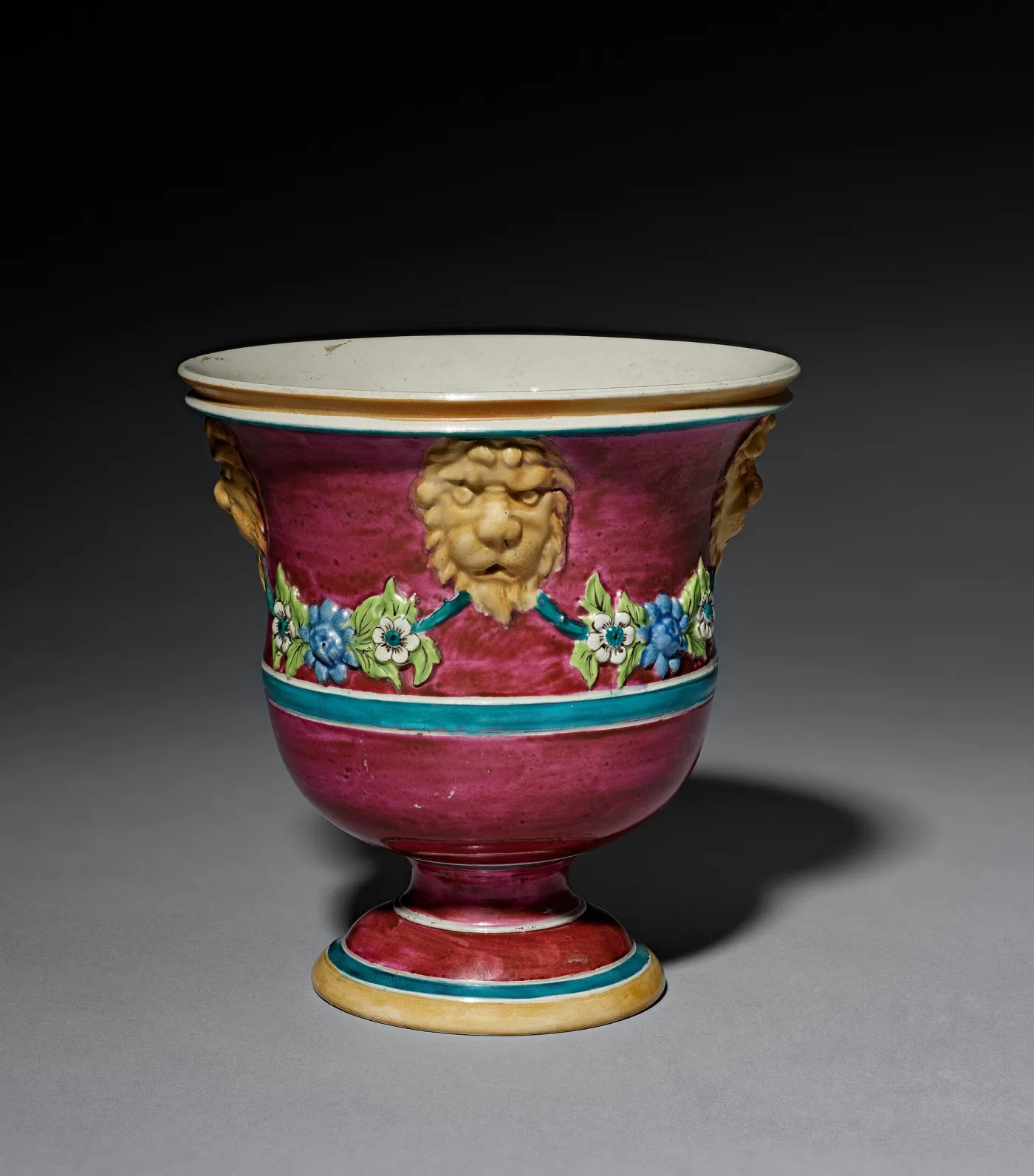Staffordshire salt-glazed jardiniere of about 1760, crimson ground with enamelled flower swags hanging from two gilt lion masks, a turquoise band below and a spreading footed base