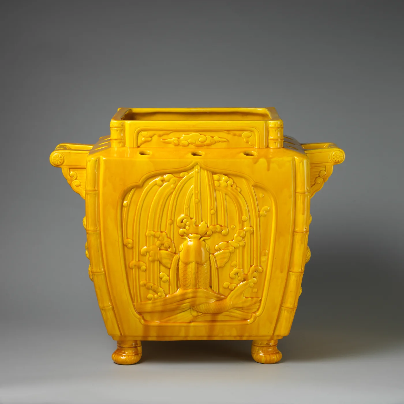 Square Minton majolica jardiniere of 1883 designed by Christopher Dresser, glazed a single deep yellow, with bamboo mouldings at the corners and rim and a moulded panel of a fountain on each face, standing on four bun feet