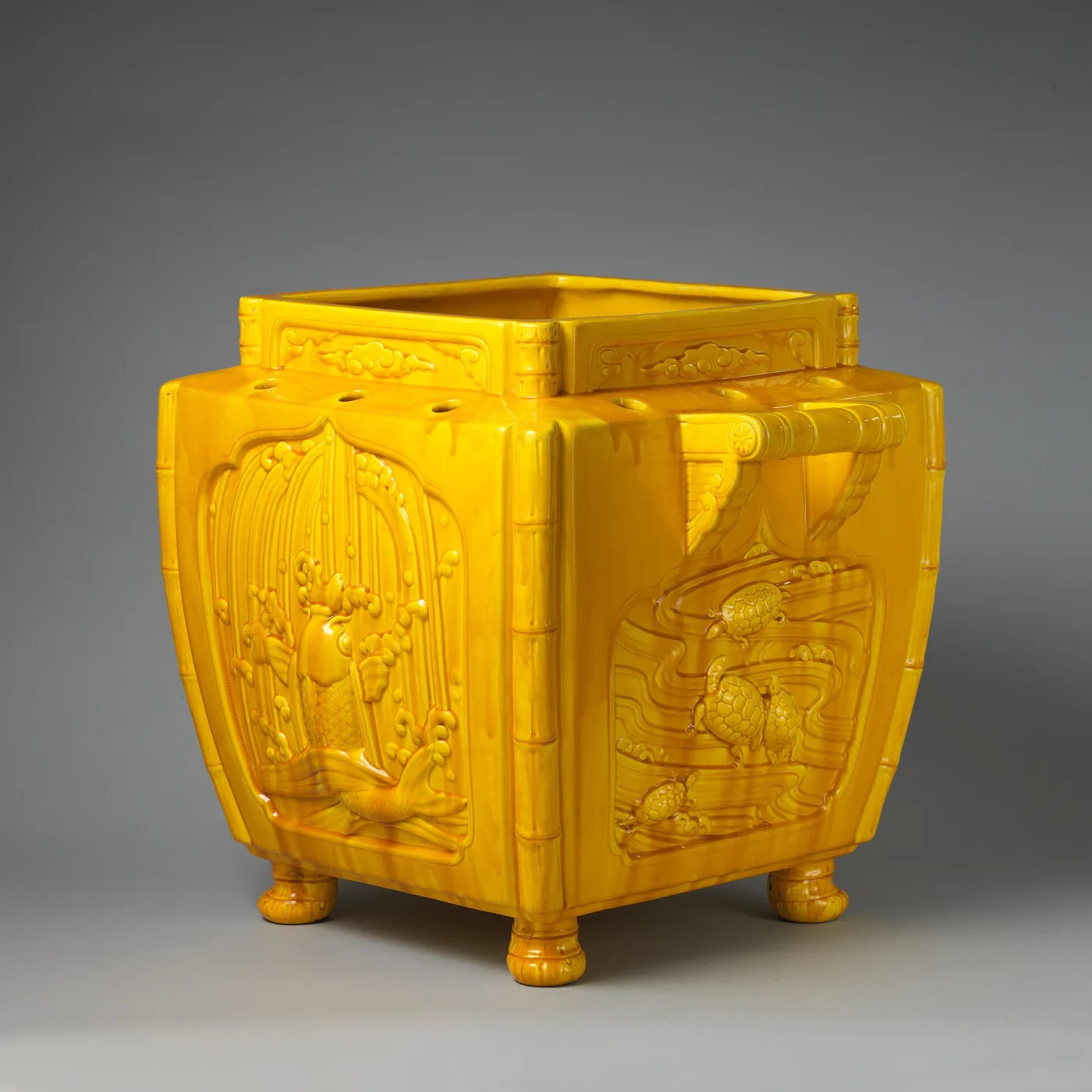 The same yellow Dresser jardiniere seen from a corner, showing that the fountain panel on one face and the panel on the adjacent face carry different moulded compositions