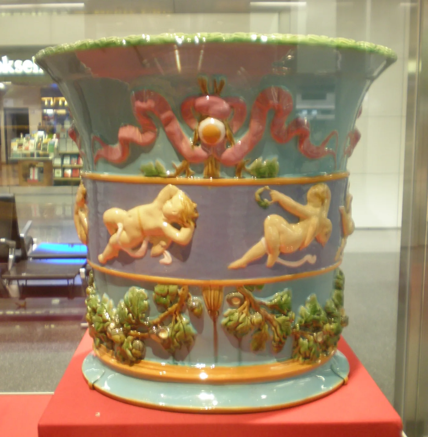 Minton majolica jardiniere of 1875 with a flaring green rim, pink ribbon swags, a sunken lavender band holding modelled nude infants in relief, and green oak leaves with brown acorns swagged below, on a turquoise foot