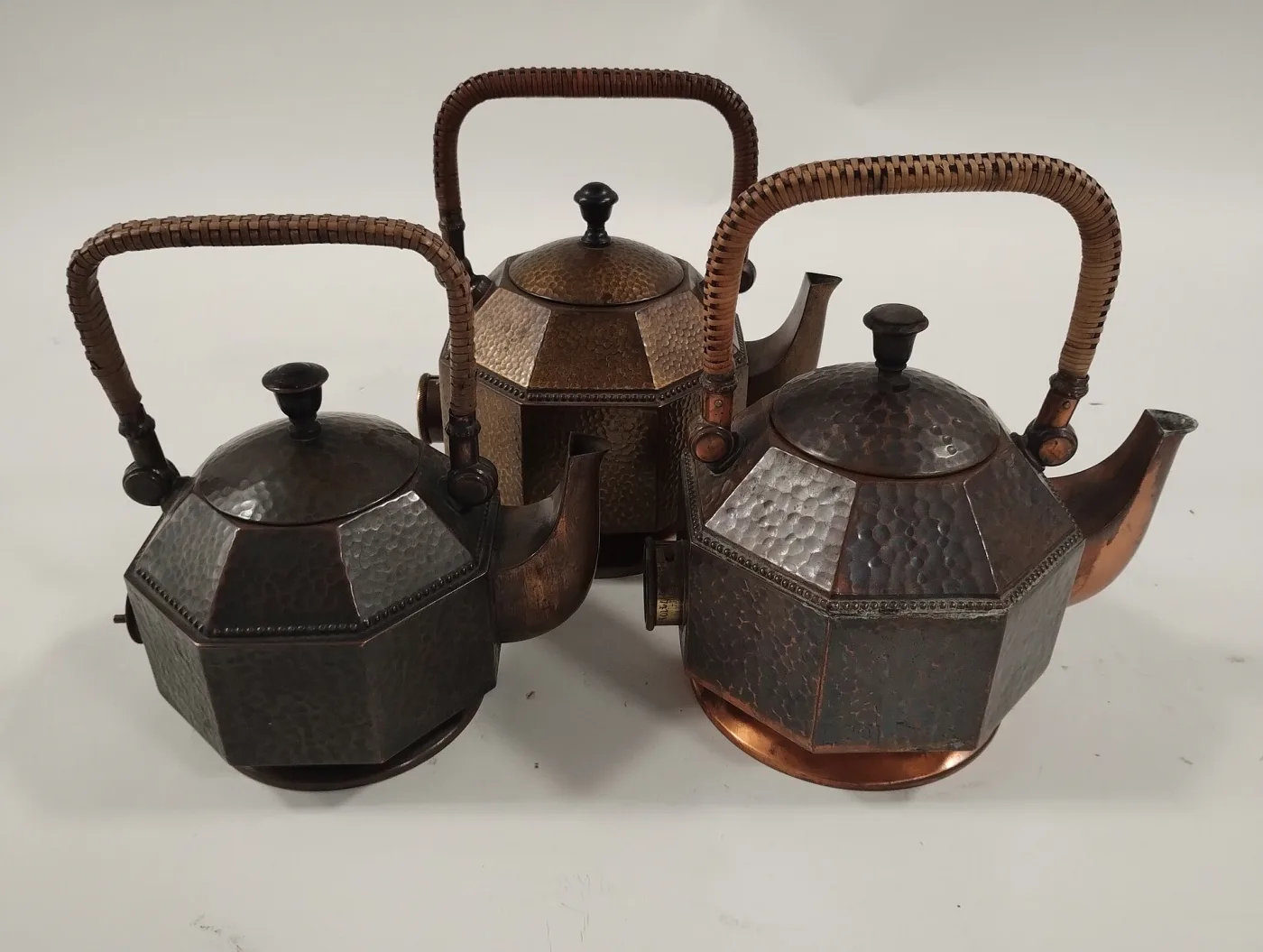 Three octagonal hammered copper kettles of increasing size, each with a rattan-wrapped bail handle, a black knob and a cylindrical fitting low on the body