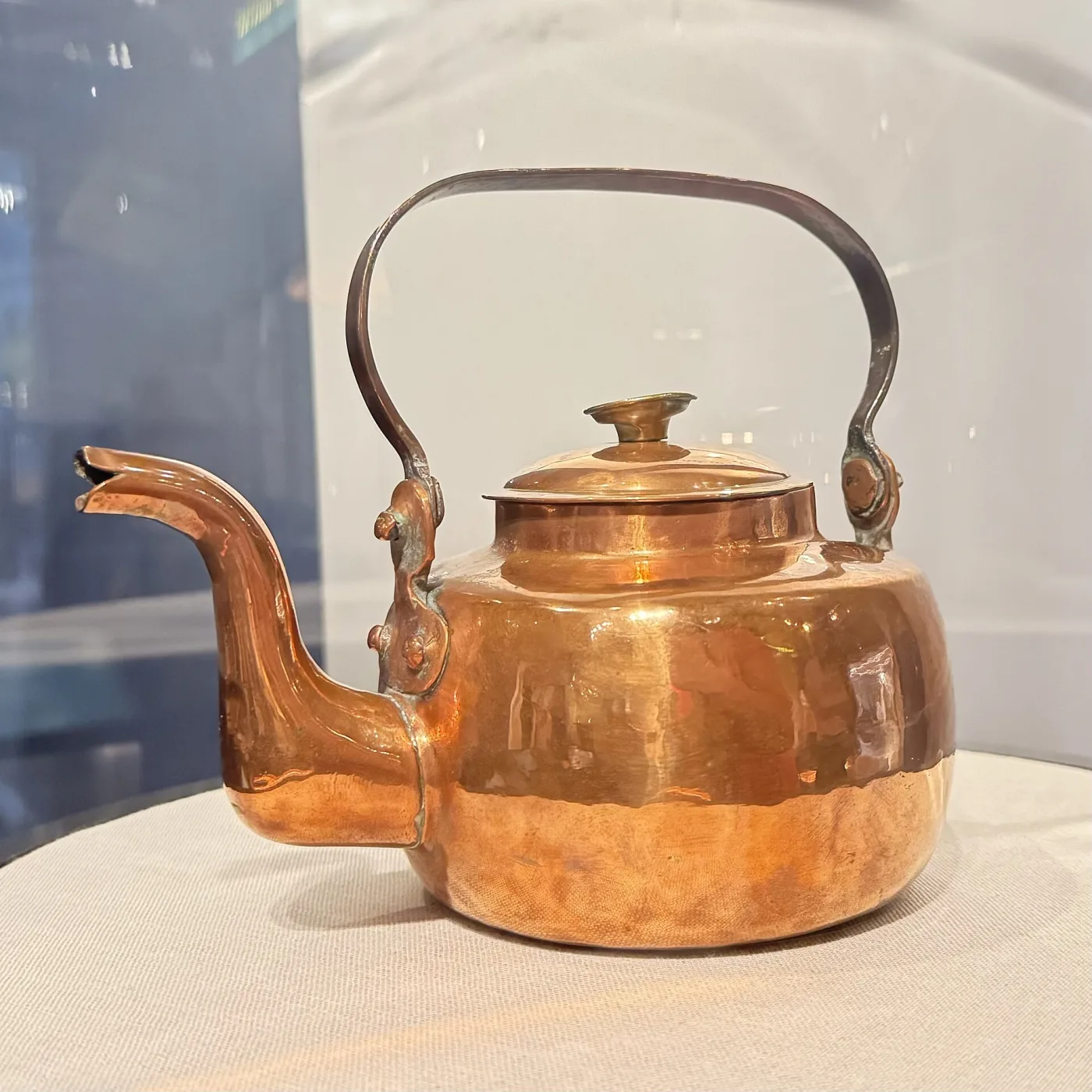 Copper kettle photographed side-on in a display case, showing a vertical seam running down the body behind the spout and cast lugs holding the swing handle
