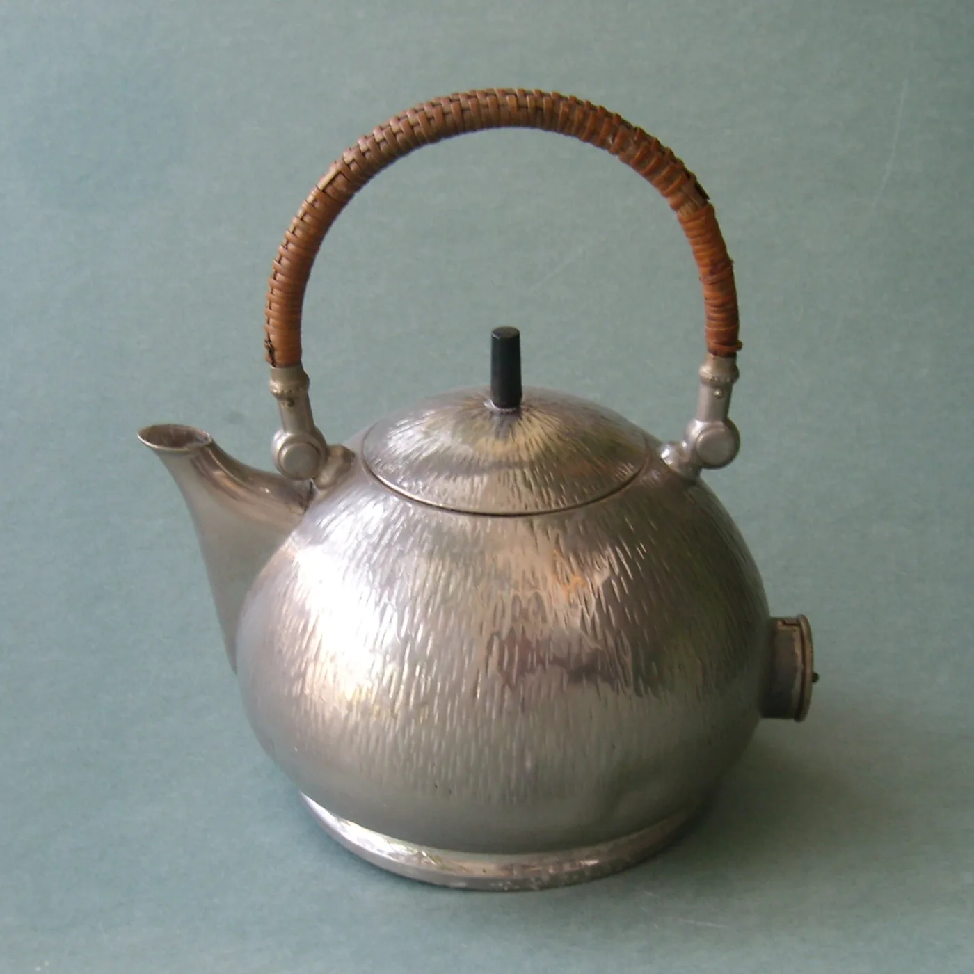 Rounded nickel-plated kettle with a finely chased vertical texture, rattan-wrapped handle and the same cylindrical fitting on the side