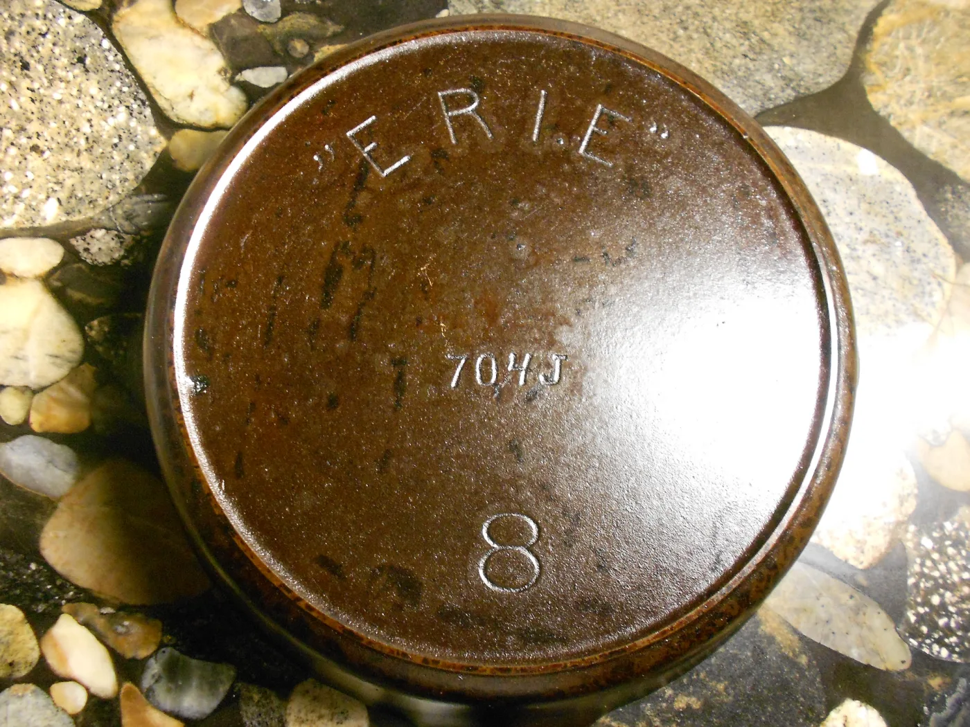 Underside of a cast-iron skillet resting on stone, its heat ring and base marked with the word ERIE in an arc above a pattern number 704J and the size numeral 8