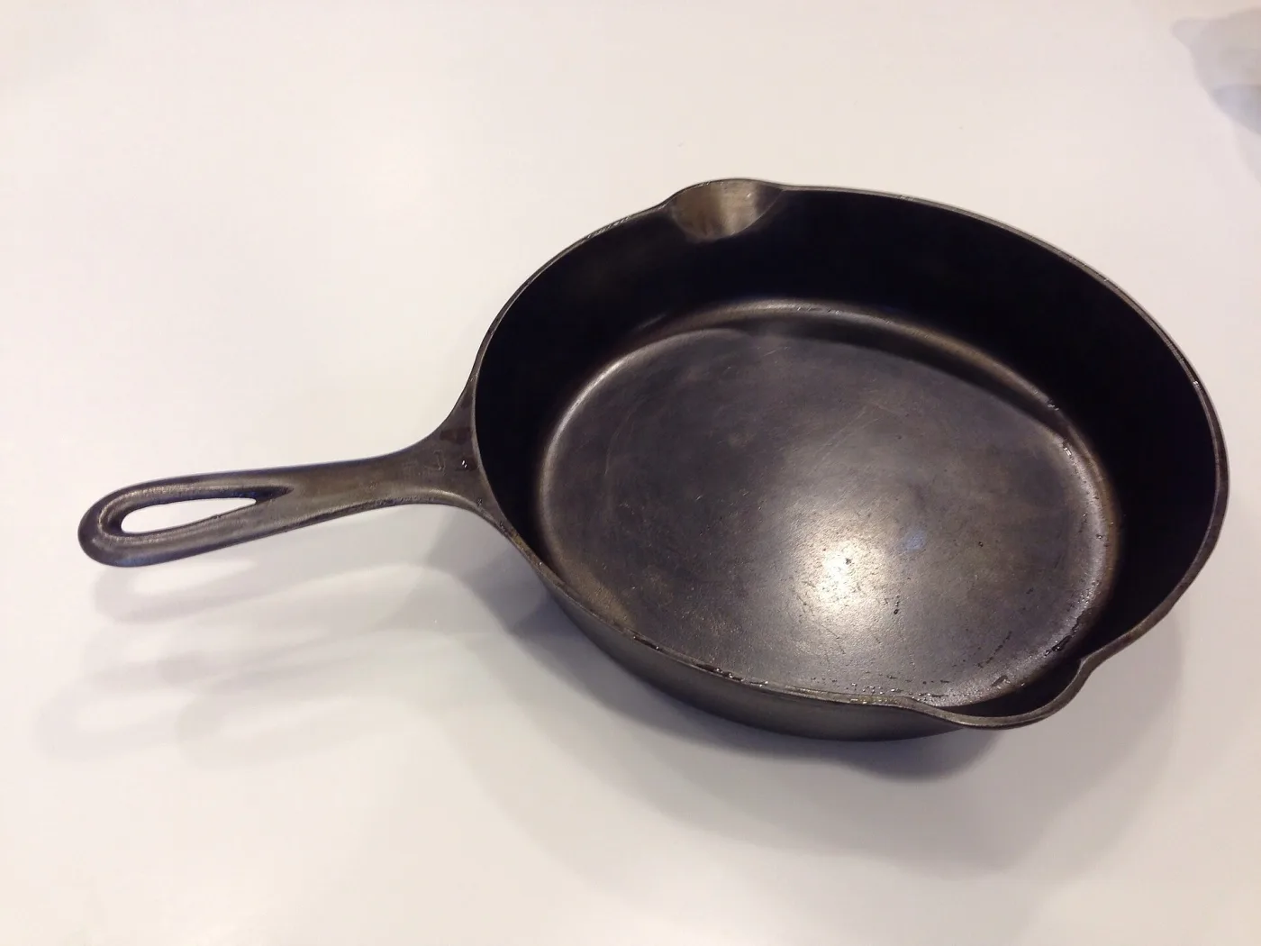 American-style cast-iron skillet seen from above, its cooking surface smooth and dark with a pouring lip either side and a long handle with a hanging hole at the end