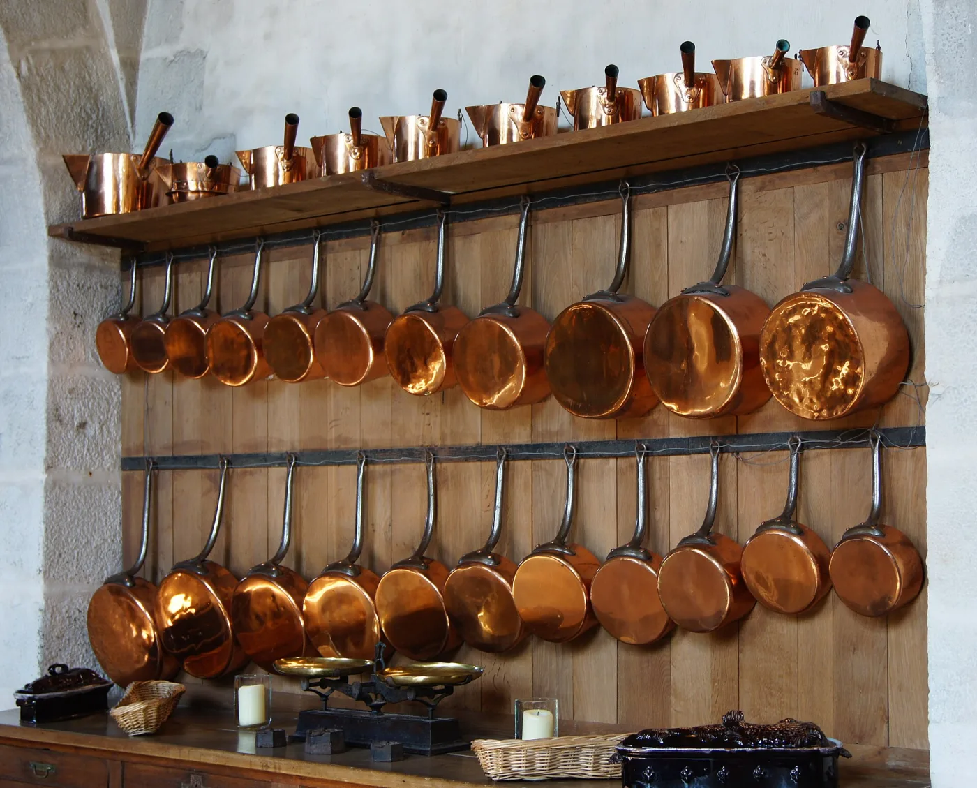Long wall rack in a chateau kitchen hung with dozens of copper saucepans, pots and moulds graded from small to large, their tinned interiors pale against polished copper exteriors