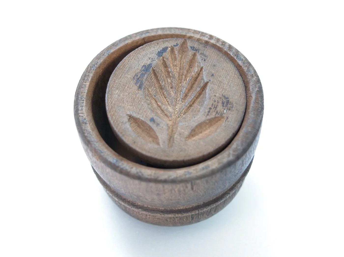 Round wooden butter stamp resting face up on a white ground, its underside carved with a stylised wheat ear inside a beaded border, the turned handle rising behind it