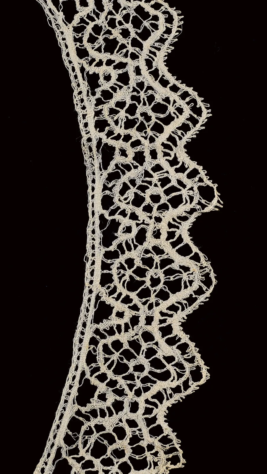 Long strip of cream bobbin lace photographed against black, its scalloped edge filled with dense floral motifs joined across an open mesh ground of regular hexagonal holes