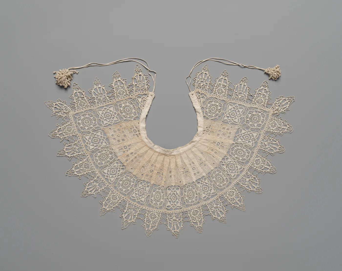 Wide semicircular lace collar of cream needle lace with a deeply pointed scalloped edge, laid flat, with a cord and two tassels attached at the neck opening